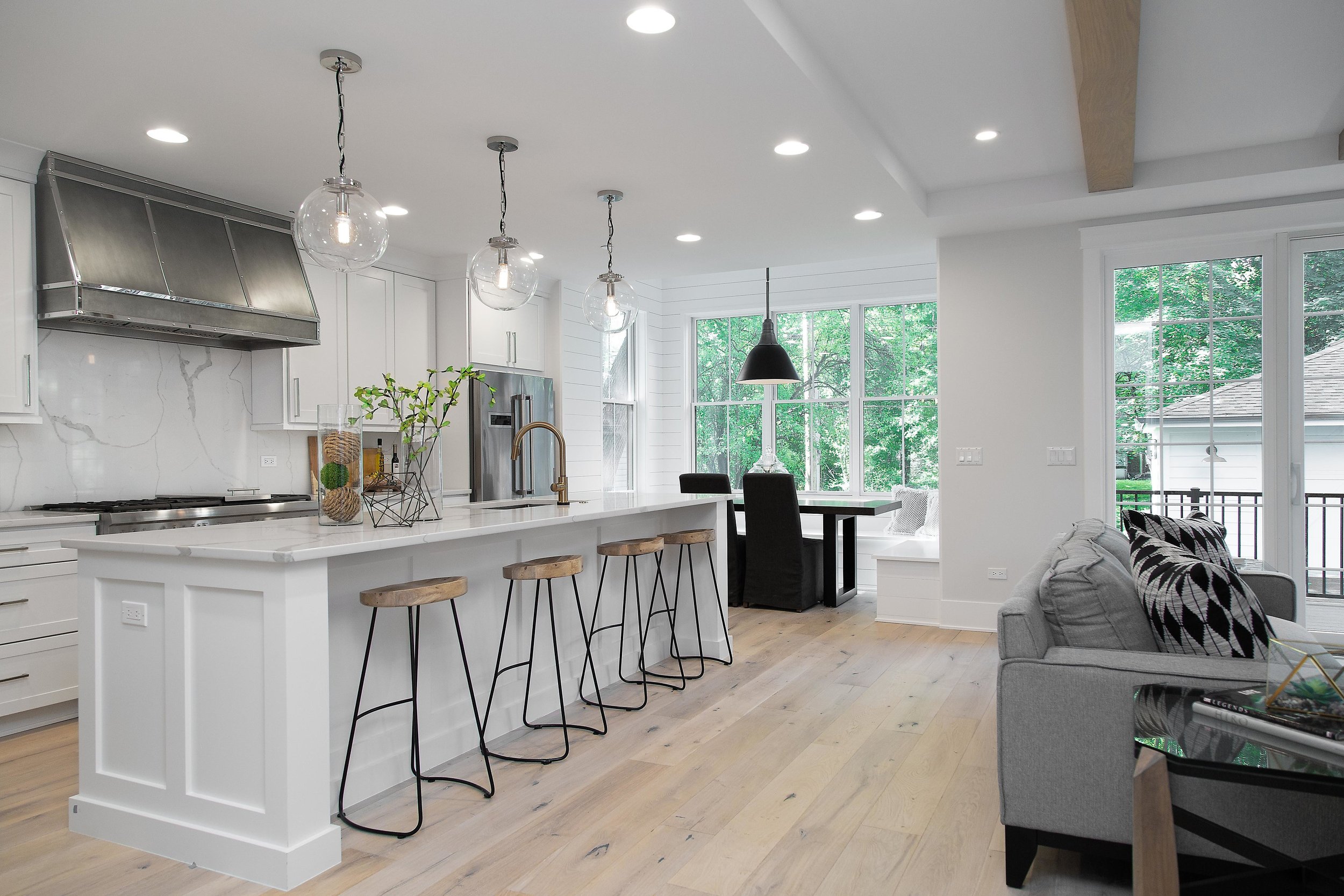 Modern kitchen with white cabinets, a large white island with wood barstools, pendant and recessed lighting, stainless steel range hood, refrigerator, and large windows showing green trees outside.