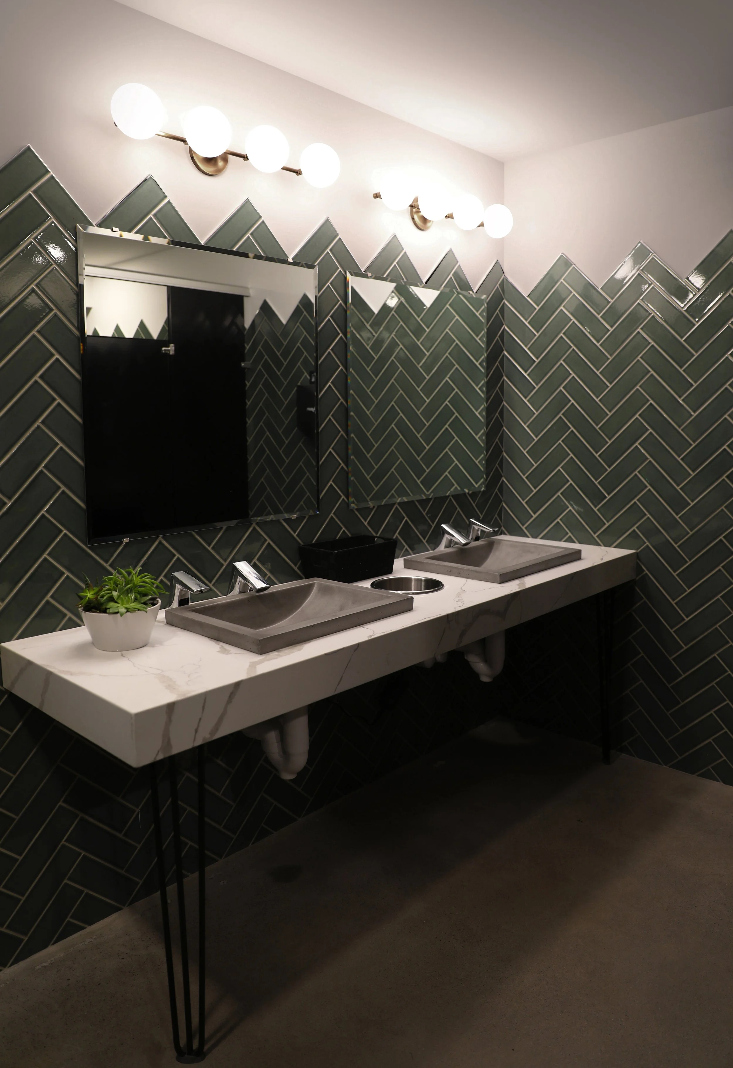 Public restroom with green herringbone tile walls, two mirrors, three sinks with modern fixtures, a plant in a white pot, and bright globe light fixtures on the ceiling.