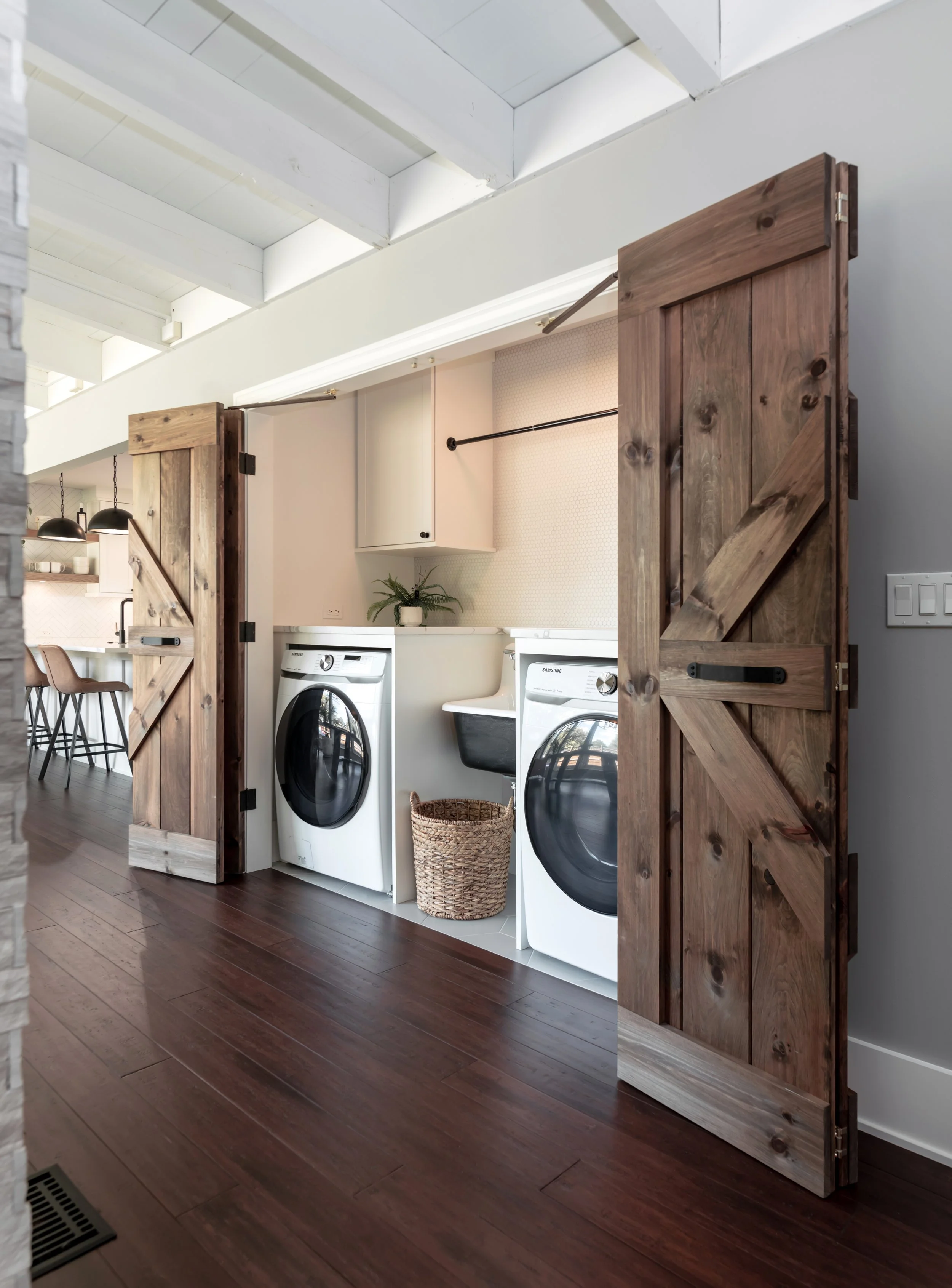 The image shows a laundry area with white washer and dryer units inside a room separated by rustic wooden sliding barn doors. There is a small basket on the floor in front of the machines and a white wall with a small shelf and a plant. Dark hardwood