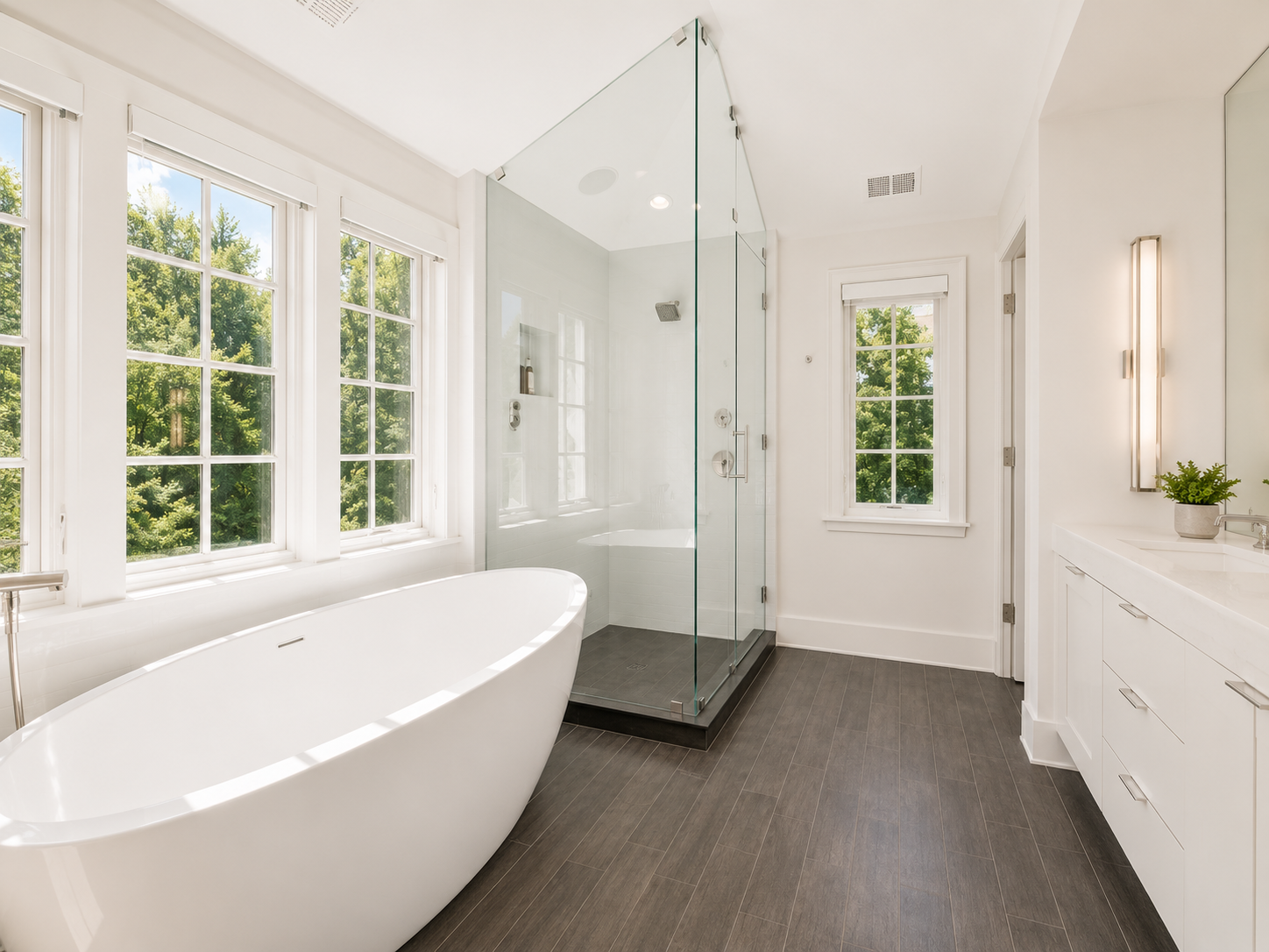 Bright modern bathroom with large windows, a white freestanding tub, a glass-enclosed shower, and white cabinetry.