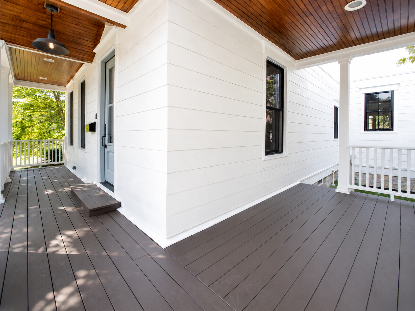 View of a porch with wood flooring, white exterior walls, black framed windows, a door, a black outdoor light fixture, a small set of steps, and a white railing. Some greenery is visible in the background.