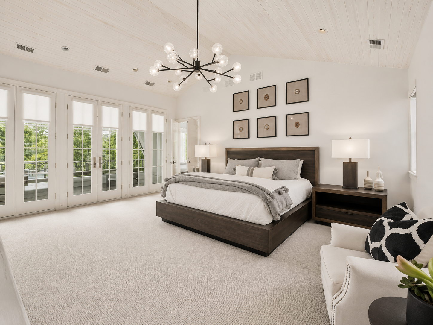 Modern bedroom with a large bed, dark wood furniture, and a beige carpet. There are nightstands with lamps, wall art above the bed, and tall glass doors leading to a balcony with greenery outside. A white armchair with a black and white pillow is in 