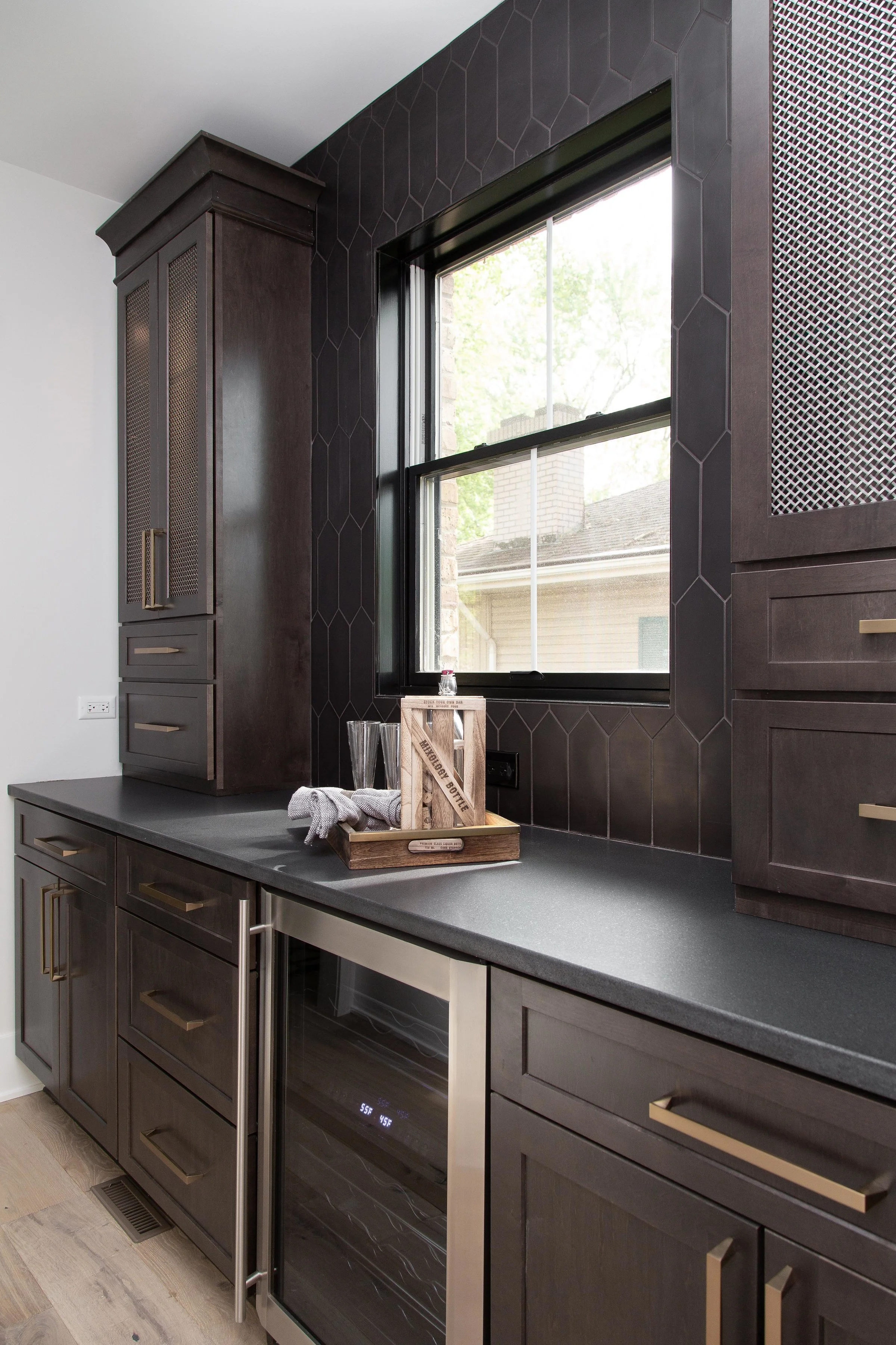 A kitchen counter with dark gray cabinets, a built-in wine fridge, a window with black trim, and a wooden tray with glassware on the countertop.
