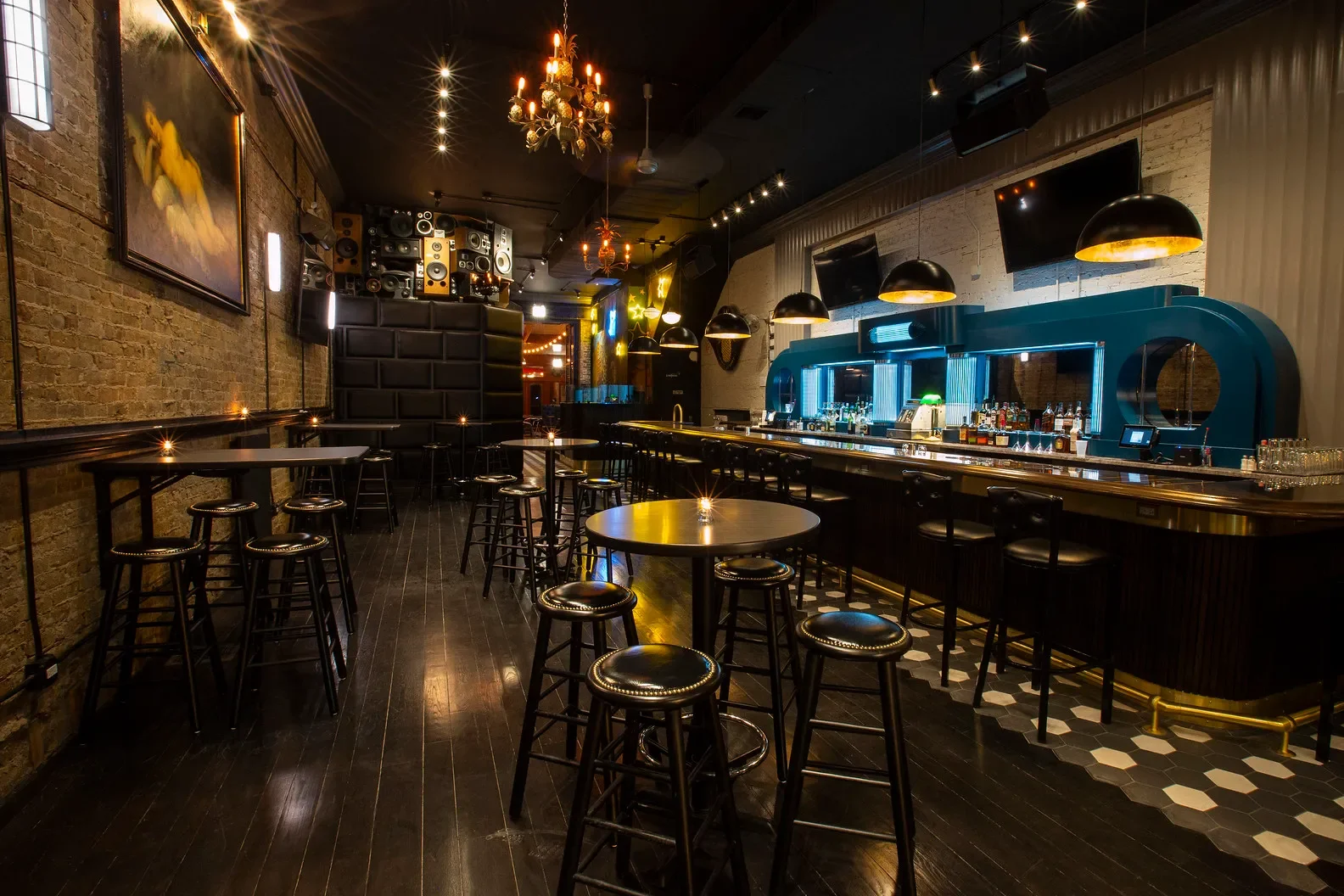 A dimly lit bar with black stools, a black bar counter with neon blue accents, hanging black pendant lights, and exposed brick walls with artwork and speakers.