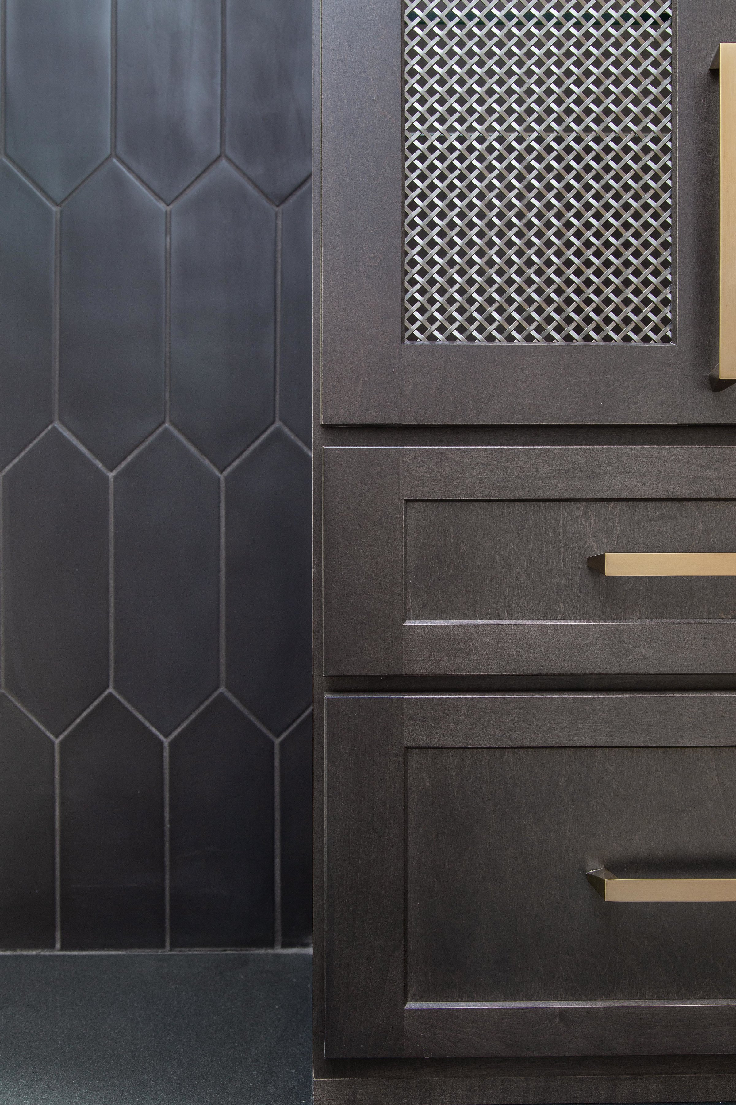 Close-up of a dark gray cabinet with gold handles and a decorative vent or grille on the door, adjacent to a wall with dark hexagonal tiles.