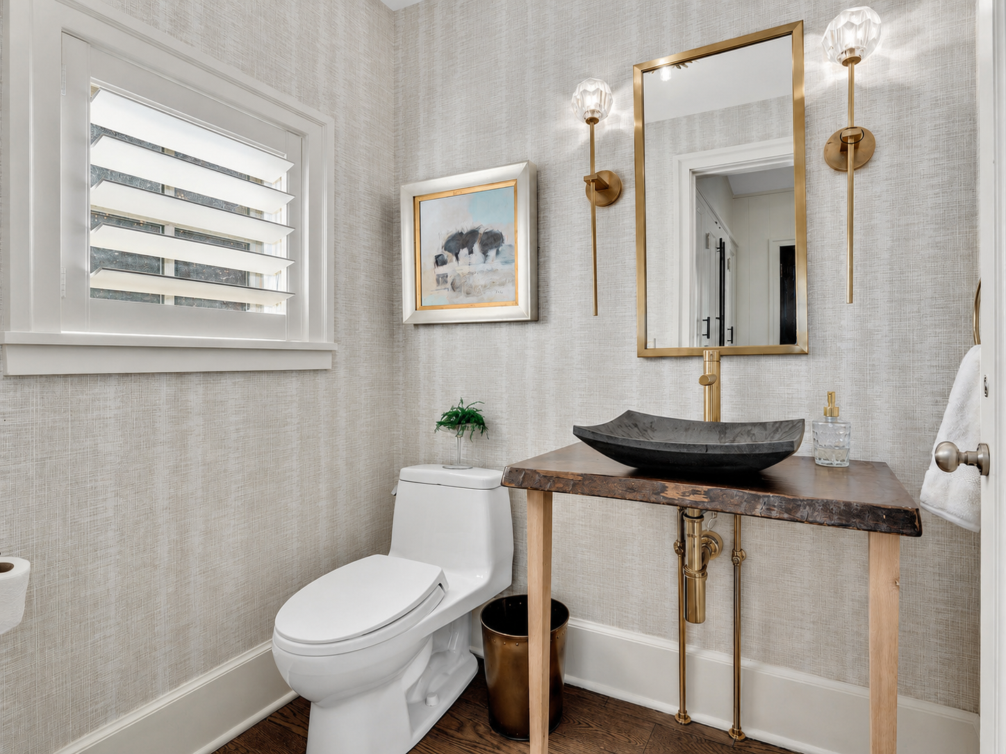 Modern bathroom with a white toilet, a wooden vanity with a black stone sink, gold fixtures, a mirror, wall sconces, a window with plantation shutters, and artwork on textured wallpaper.