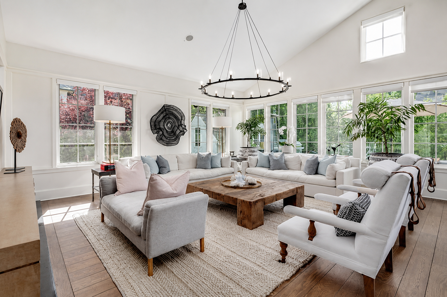 Bright living room with white walls and large windows, featuring a central rustic wooden coffee table, white sofas with pastel pillows, and a chandelier above.