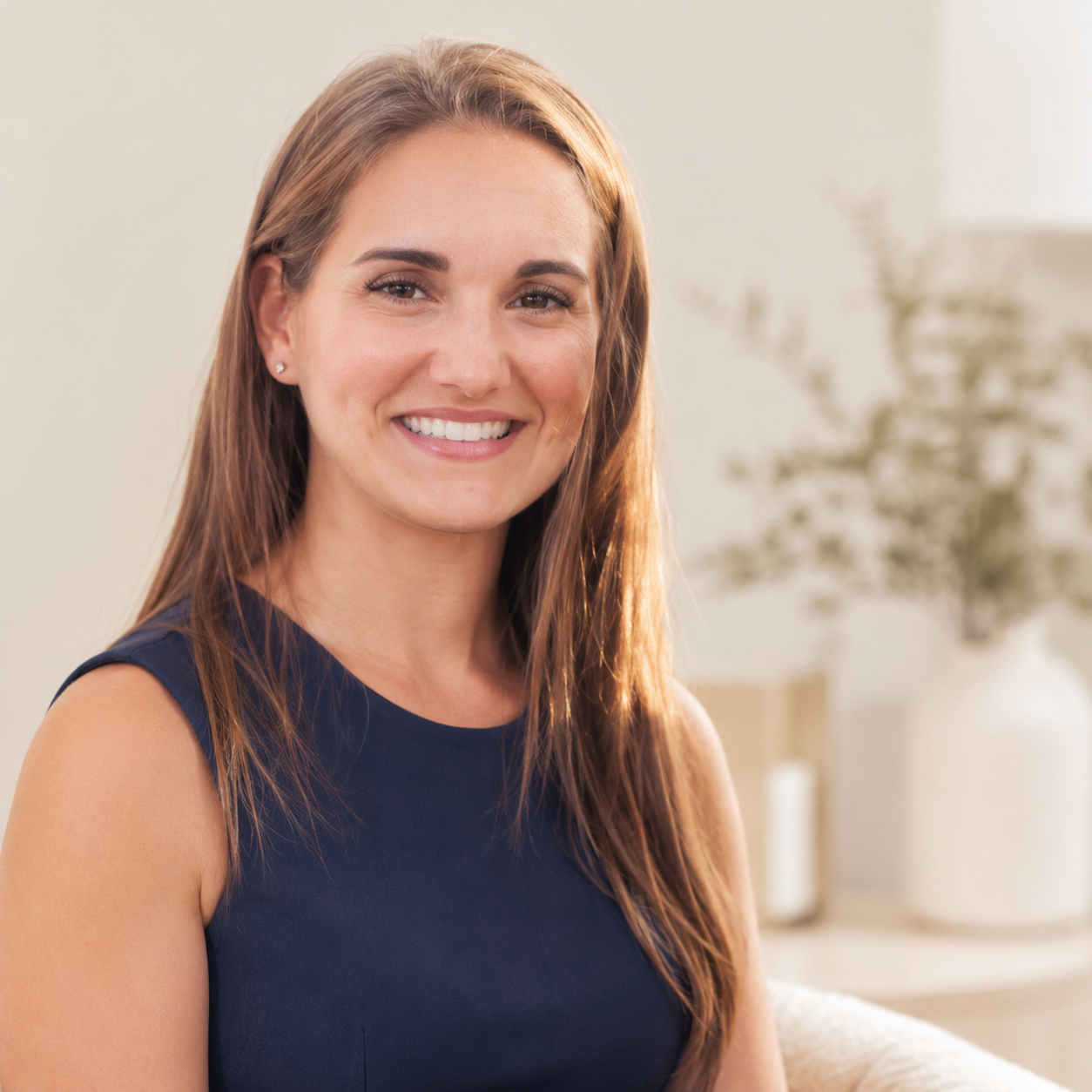 Woman with long brown hair smiling and wearing a navy sleeveless top, sitting in a bright room with a blurred background including a plant and a cream-colored wall.
