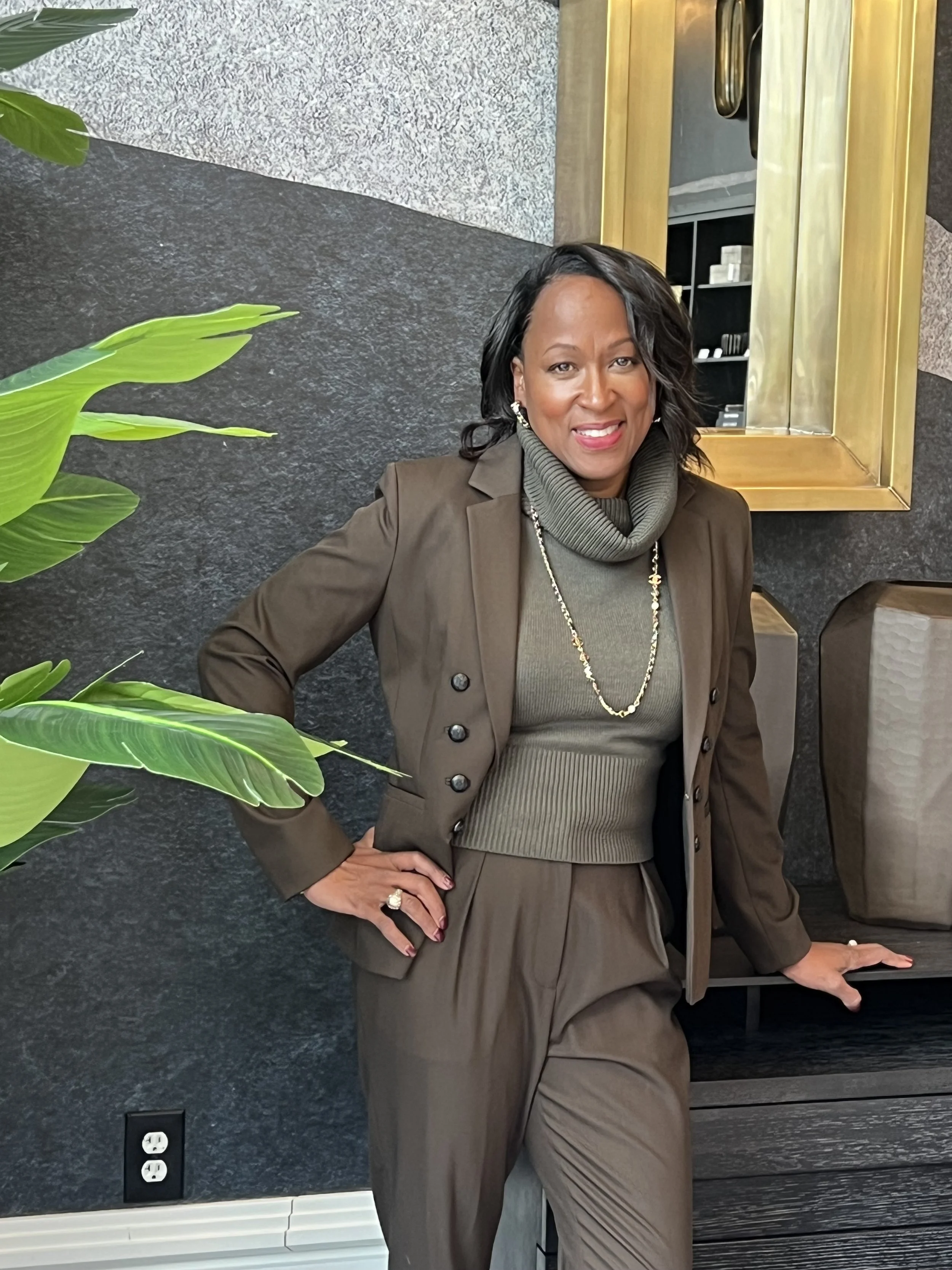 A woman with dark curly hair wearing a brown blazer, beige turtleneck sweater, and matching brown pants, standing indoors against a black textured wall with a large gold-framed mirror behind her, and green plant leaves partially visible on the left.
