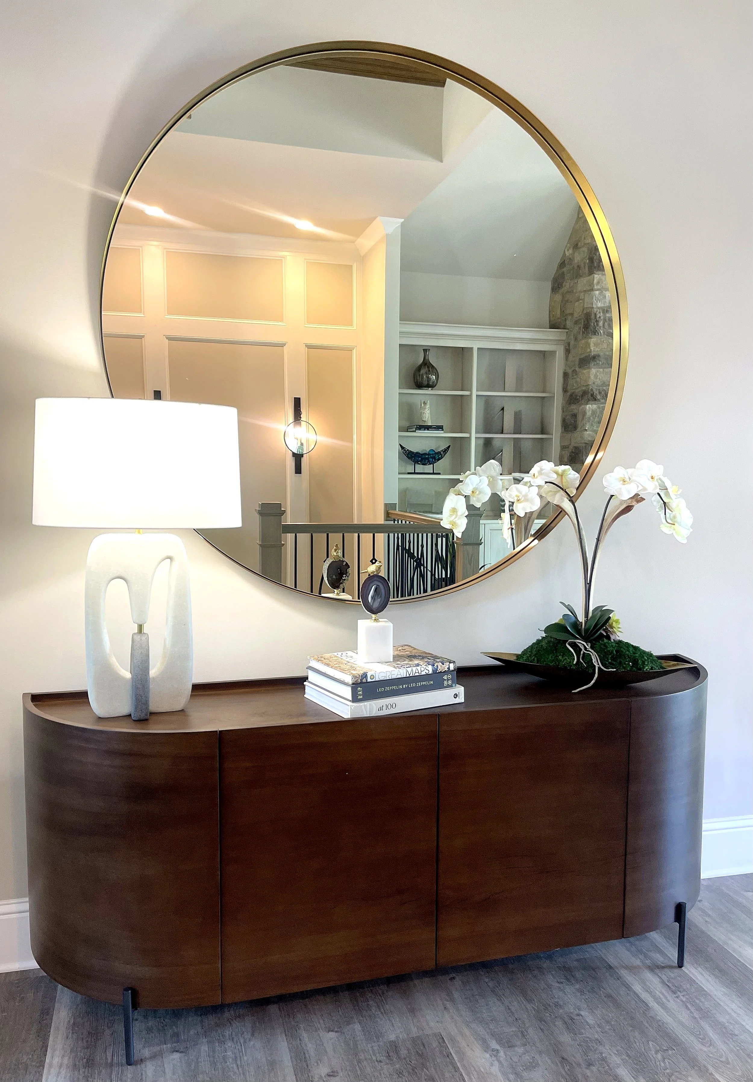 A dark wood sideboard with a curved front positioned against a white wall. On top of the sideboard is a white modern ceramic table lamp, a stack of books, and a decorative piece. A large round mirror with a gold frame hangs on the wall behind, reflecting part of a room with a nook, bookshelf, and stone fireplace. There is also a white orchid plant in a black tray on the sideboard.