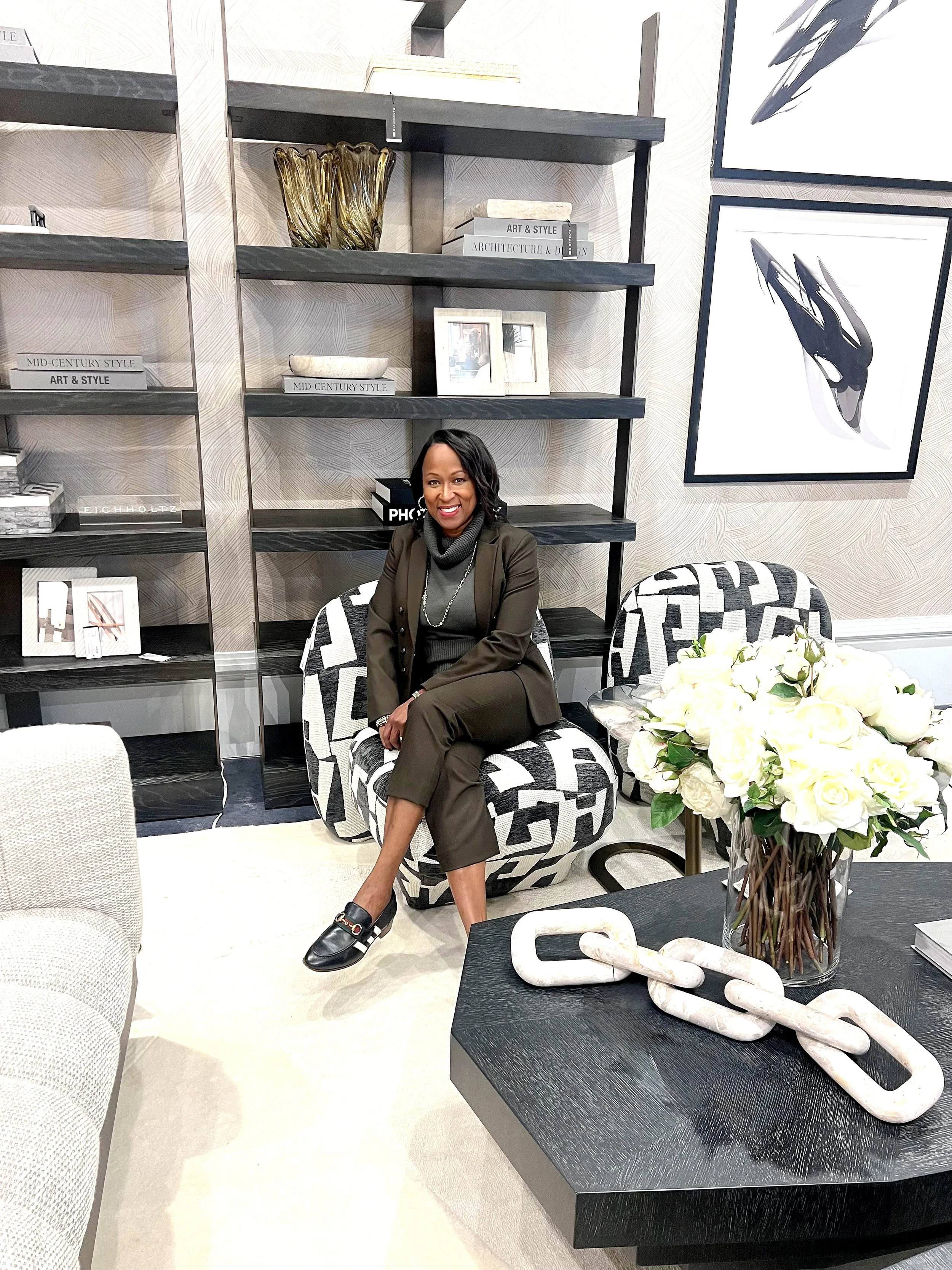 A woman sitting in a modern, stylish living room with a black and white patterned chair, a bouquet of white roses on a black table, and decorative books and art on black shelves behind her. She is smiling, dressed in dark green pants and a blazer.