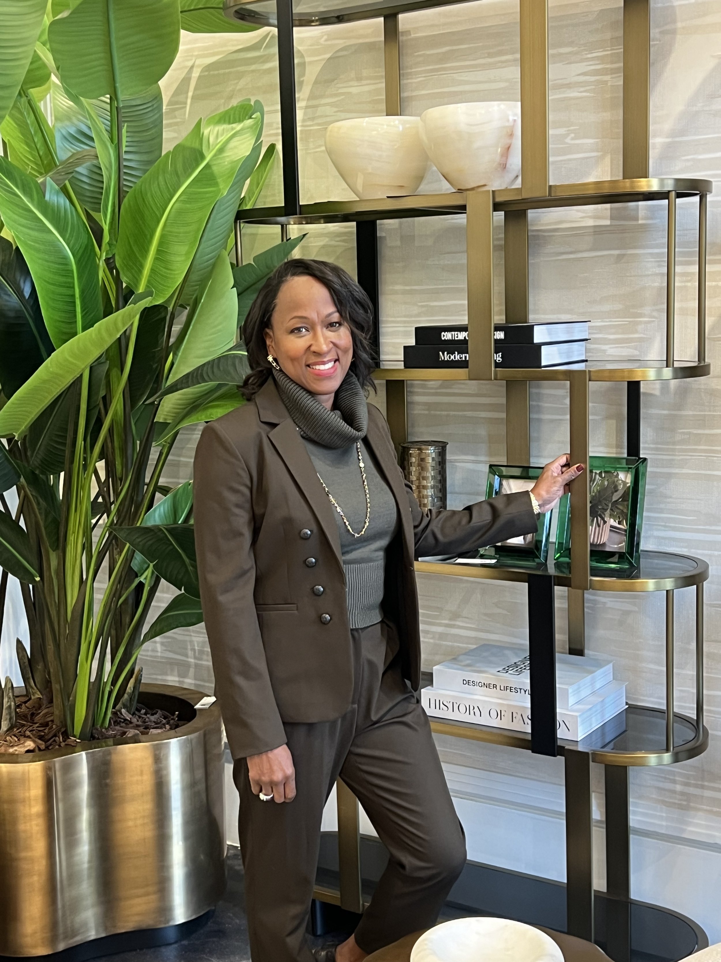 A woman with dark hair, wearing a brown suit and gray turtleneck and jewelry, standing next to a large leafy plant and decorative shelves with books and bowls in a modern interior.