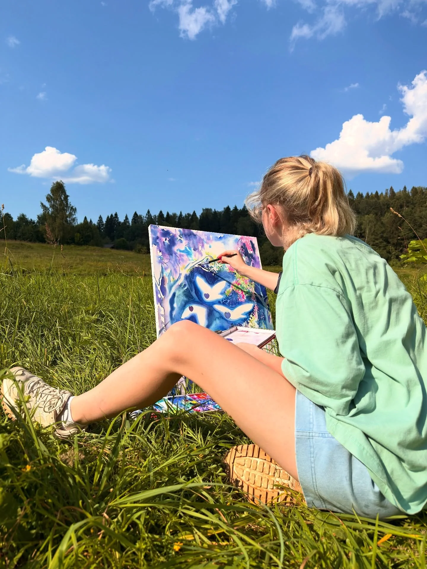 A girl with blonde hair tied in a ponytail, wearing a green shirt and blue shorts, sitting in a grassy field, painting on a canvas with a vibrant, abstract landscape, under a clear blue sky with scattered clouds, surrounded by trees.