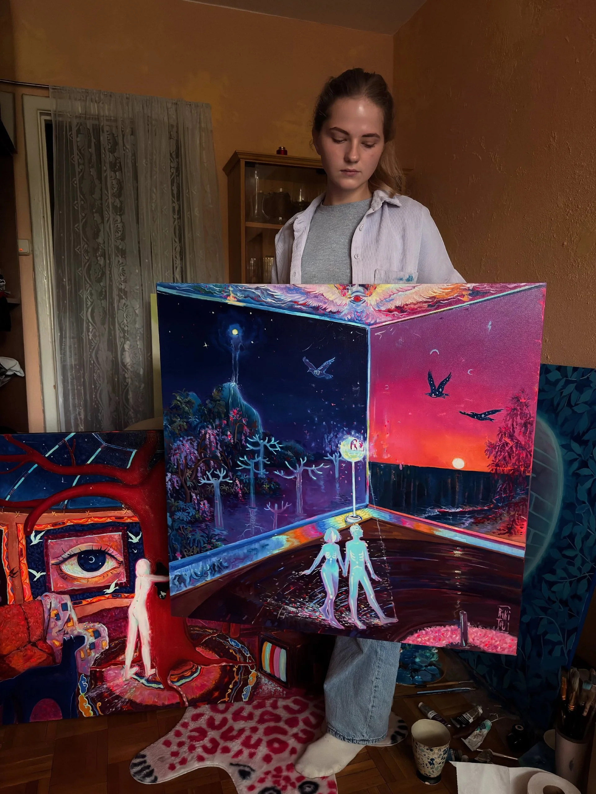 An artist standing in a room holds a large, colorful painted canvas. The painting depicts a surreal scene with two figures holding hands under a vibrant pink and purple sky with flying birds. The room has warm-toned walls, a wooden cabinet, and various art supplies and paintings visible around.