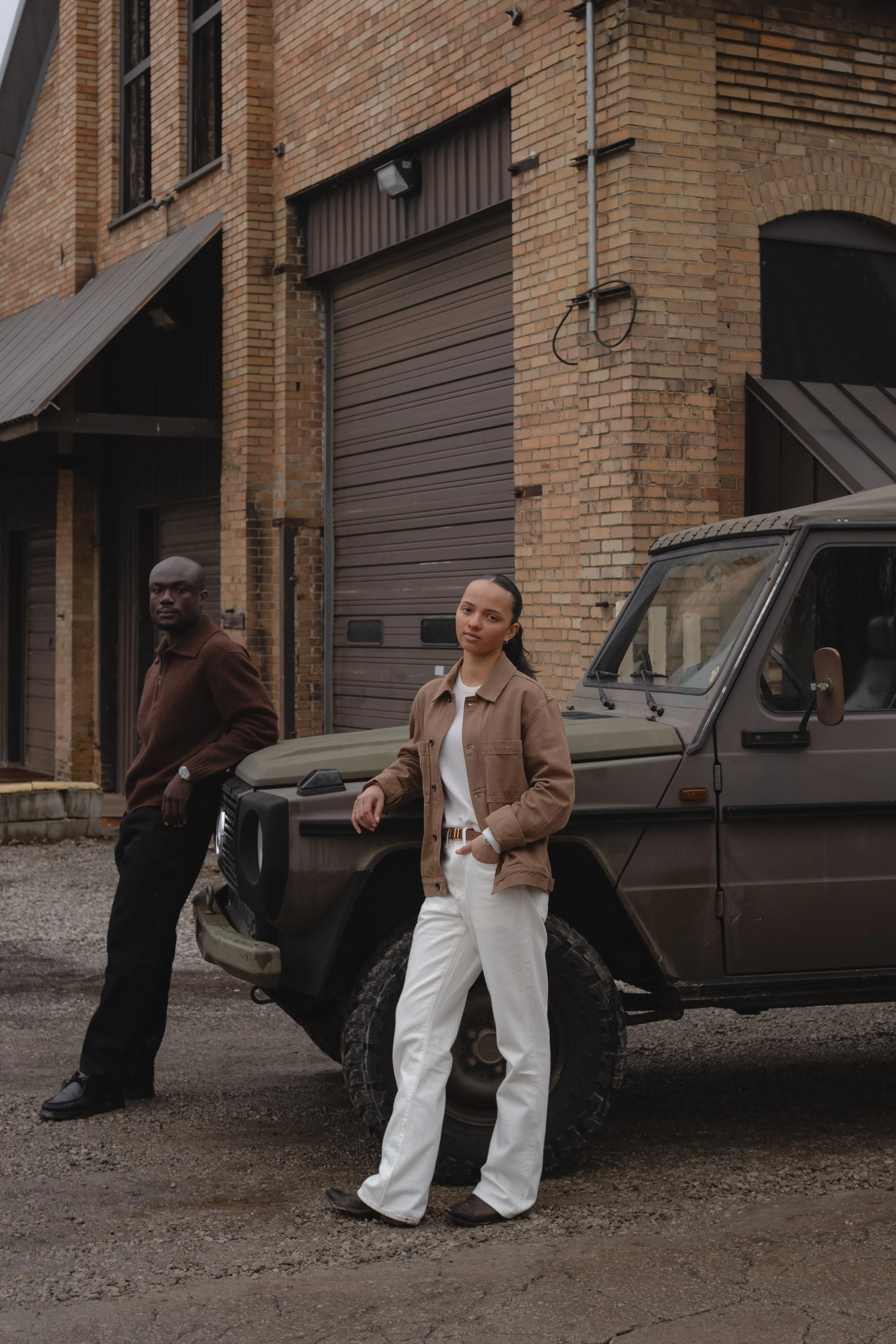 A man and a woman standing in front of a dark green vehicle on a gravel street, with a brick building in the background.