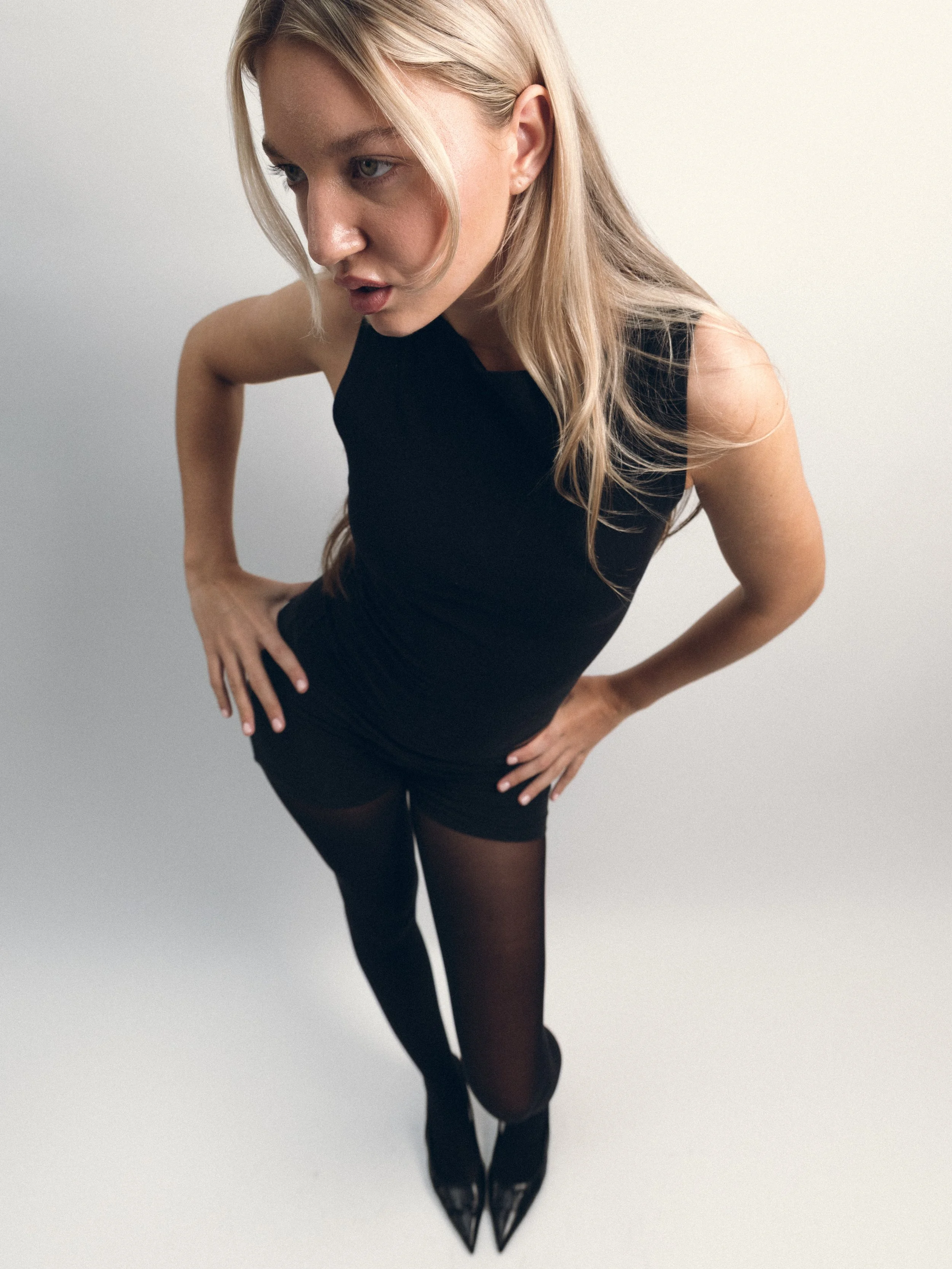 A woman with blonde hair wearing a black dress, tights, and heels, standing with hands on her hips against a plain white background.