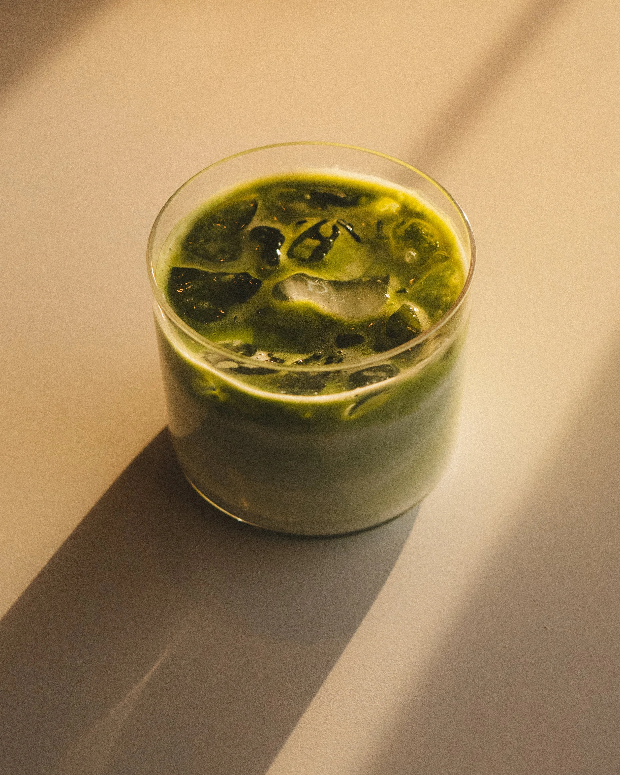 A clear glass with a greenish liquid containing dark-colored ingredients, placed on a white surface with a shadow cast to the left. Columbus, Ohio, David Kim Photography