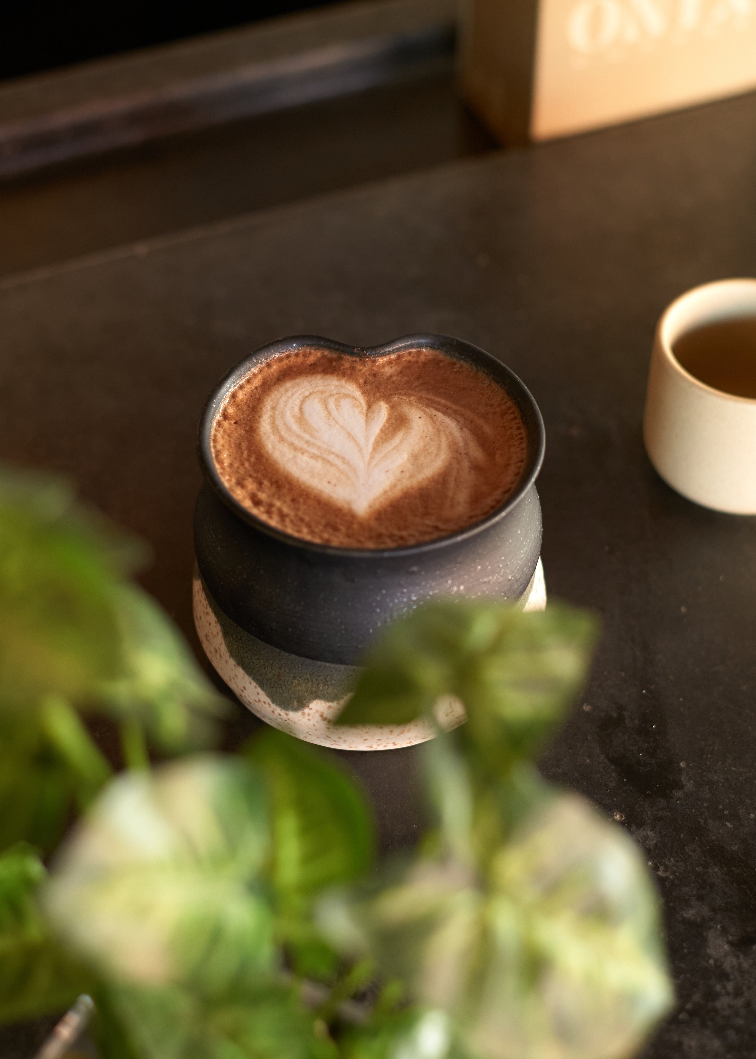 Close-up of a ceramic mug of coffee with latte art heart on a dark surface, with a white cup of tea and green leaves partially visible in the foreground. Columbus, Ohio, David Kim Photography