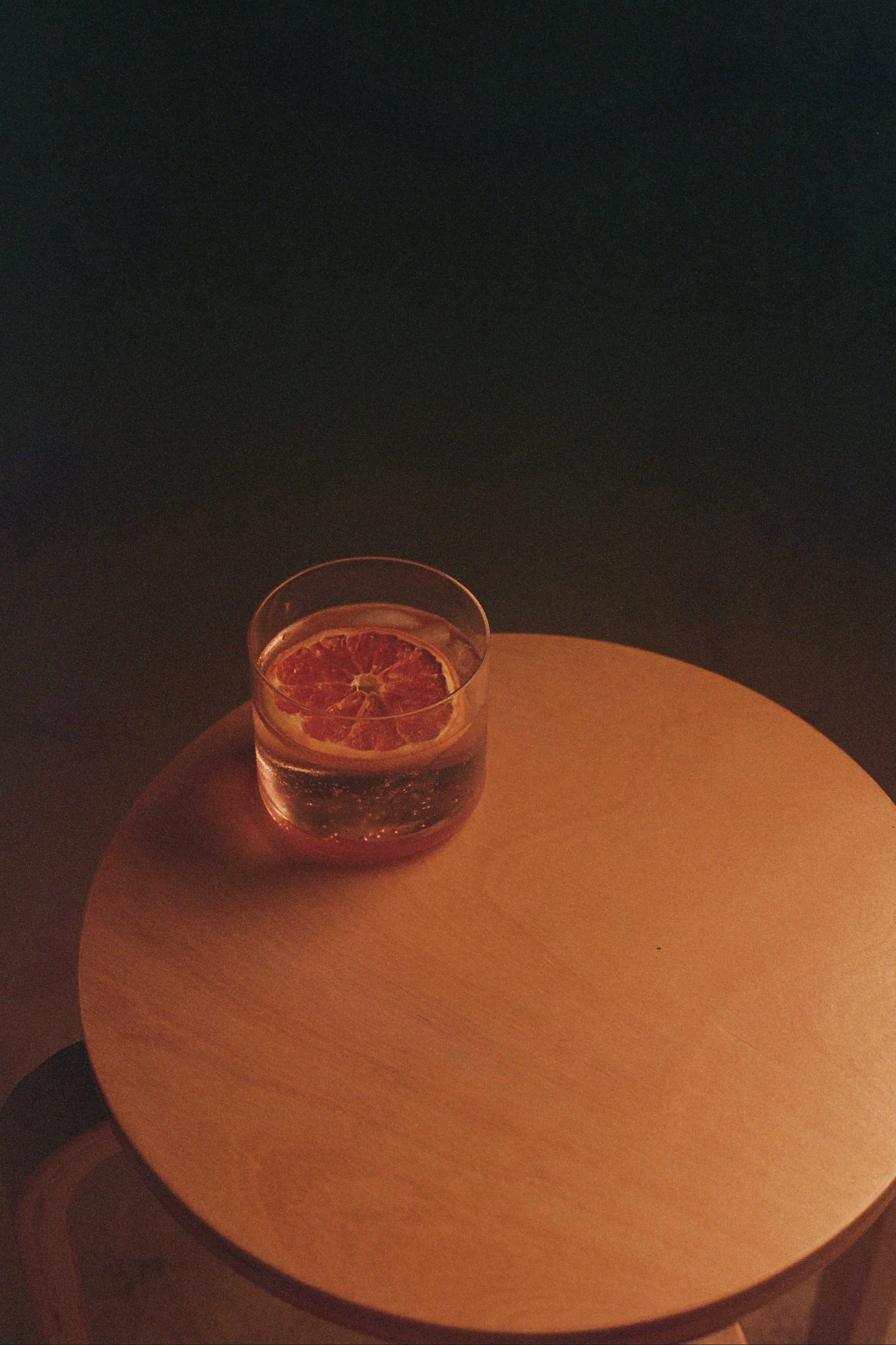 A glass of water with a slice of dried orange inside, placed on a round wooden table in a dimly lit room. Columbus, Ohio, David Kim Photography