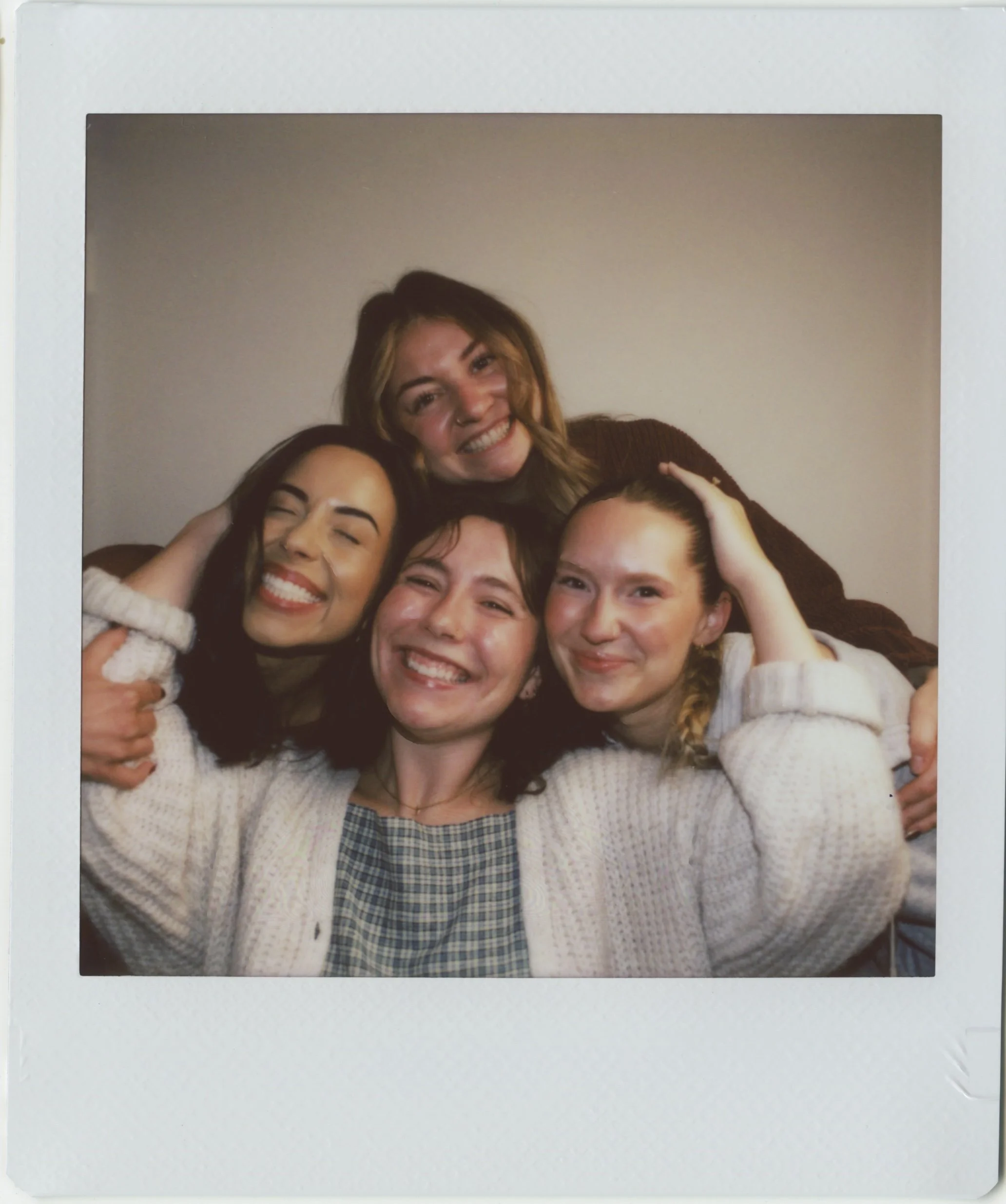 Group of smiling young women hugging and leaning on each other, taking a selfie together in a cozy indoor setting.