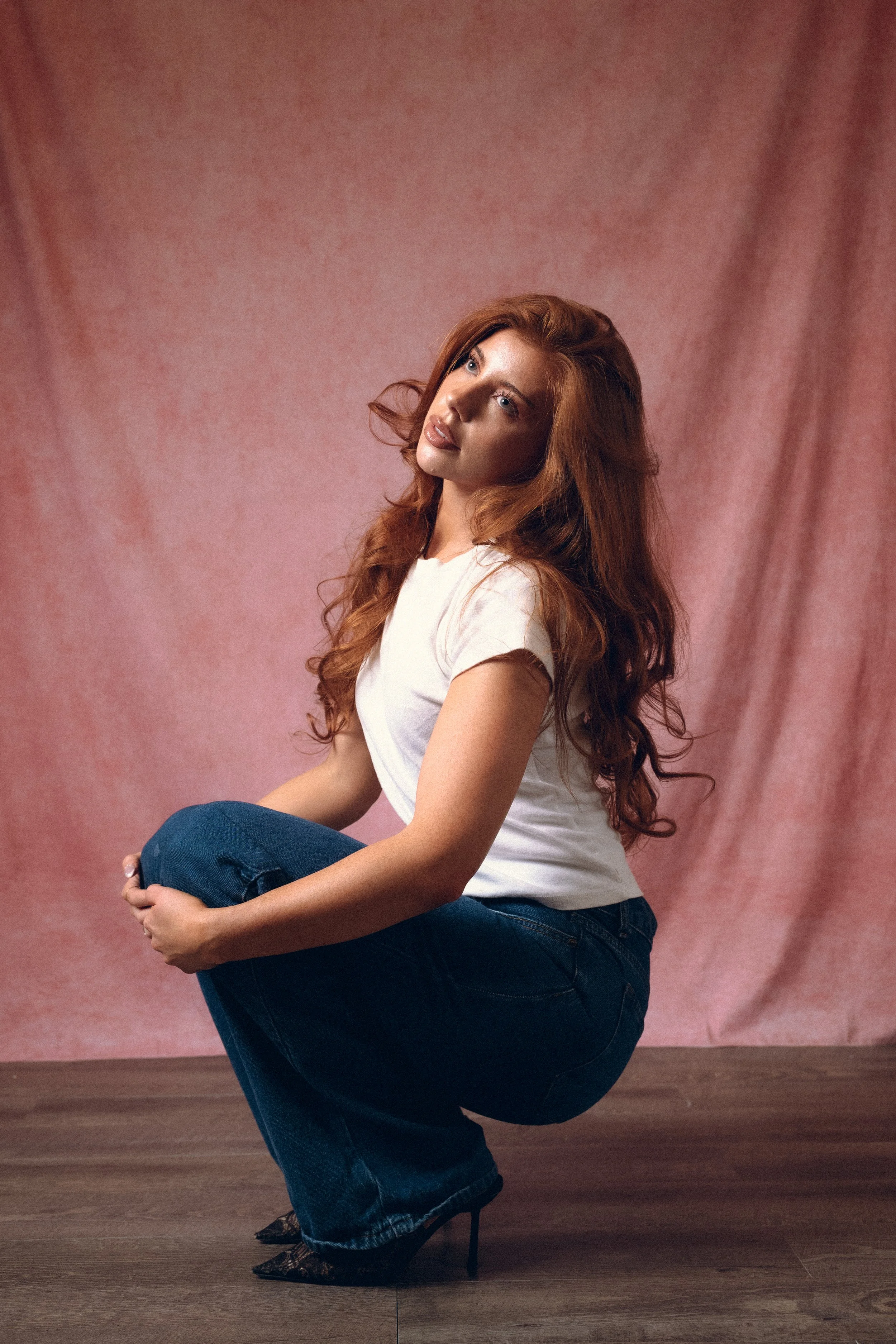 A woman with long, wavy red hair and light skin poses in front of a pink backdrop. She is wearing a white t-shirt and dark blue jeans, and is crouching with one knee up, holding her foot with her hands and wearing high heels.Columbus, Ohio, David Kim