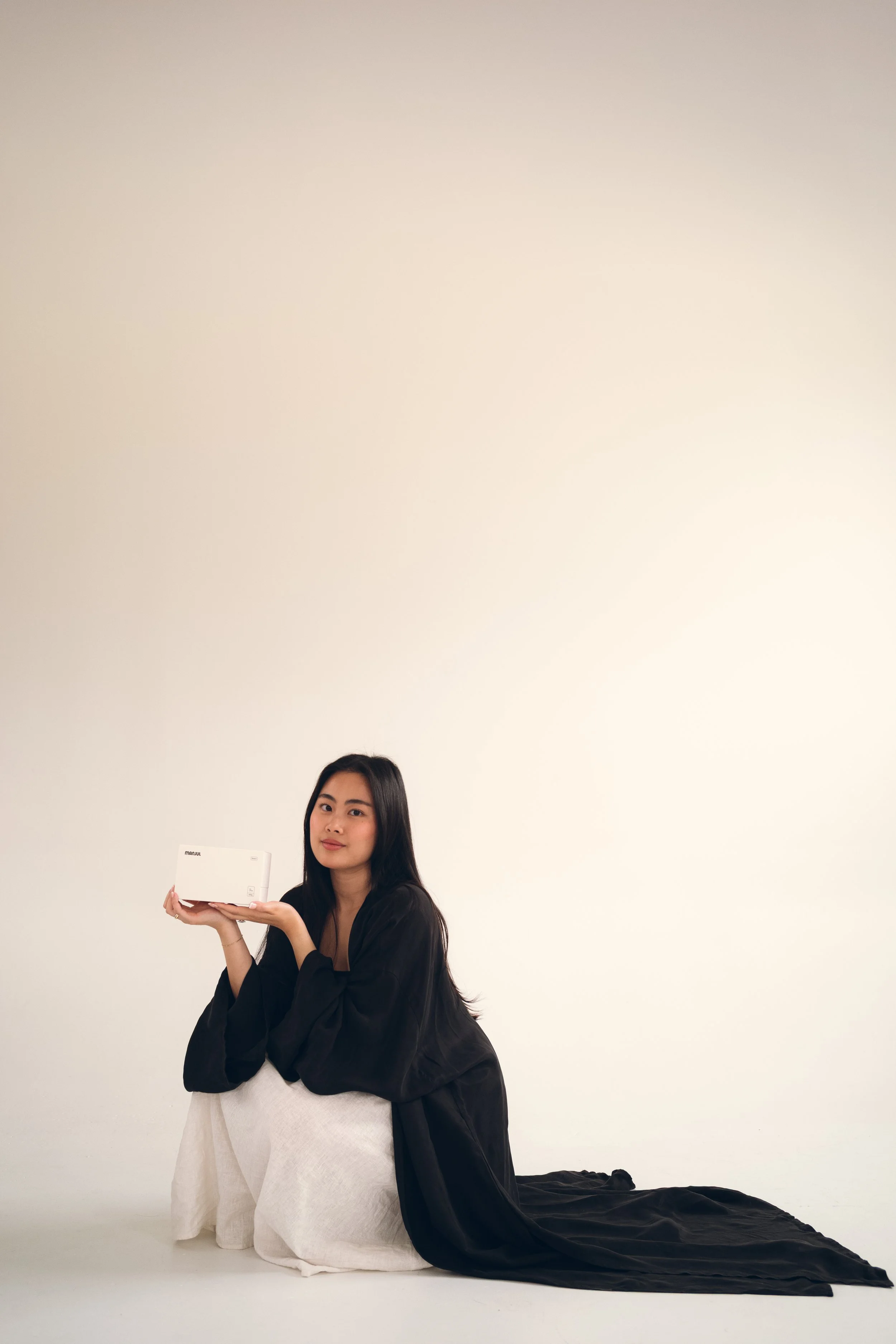 A woman with long dark hair wearing a black jacket over a white dress, sitting on the floor with her legs to the side, holding a white box, and looking at the camera against a plain white background. Columbus, Ohio, David Kim Photography