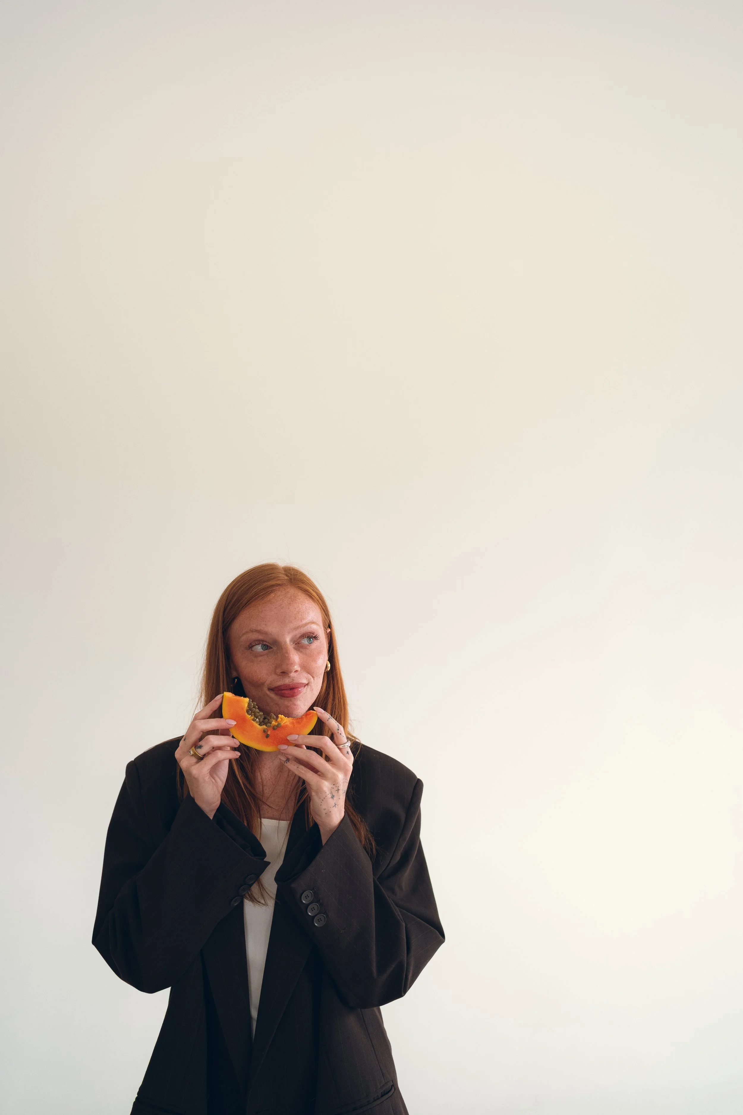 A woman with red hair and freckles wearing a black blazer is holding a slice of papaya fruit with seeds and is looking to her right against a plain light-colored background. Columbus, Ohio, David Kim Photography