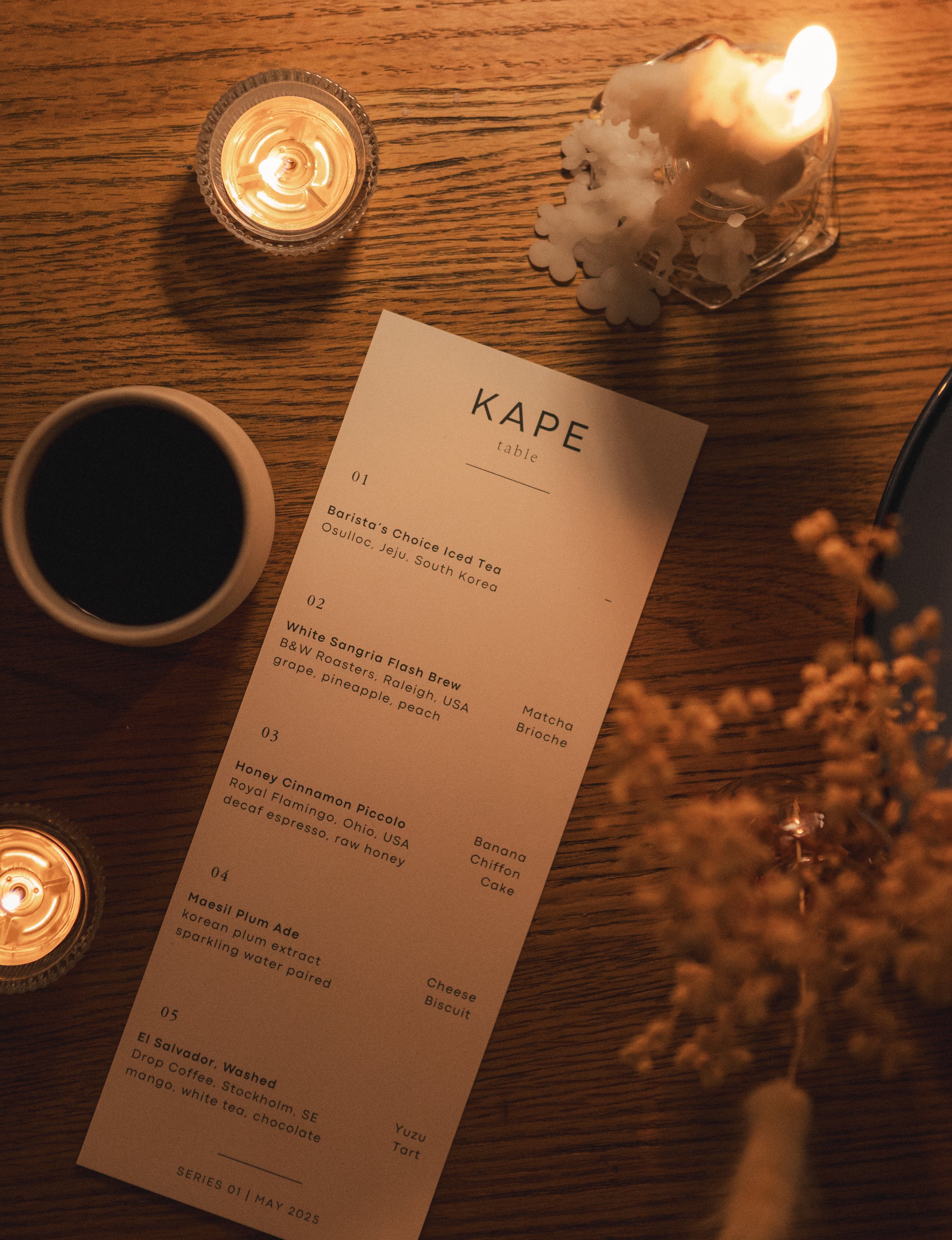 A tabletop with three lit candles, a tea menu, a cup of black coffee, a small glass jar with white flower-shaped confetti, and a few blurred flowers in the foreground.