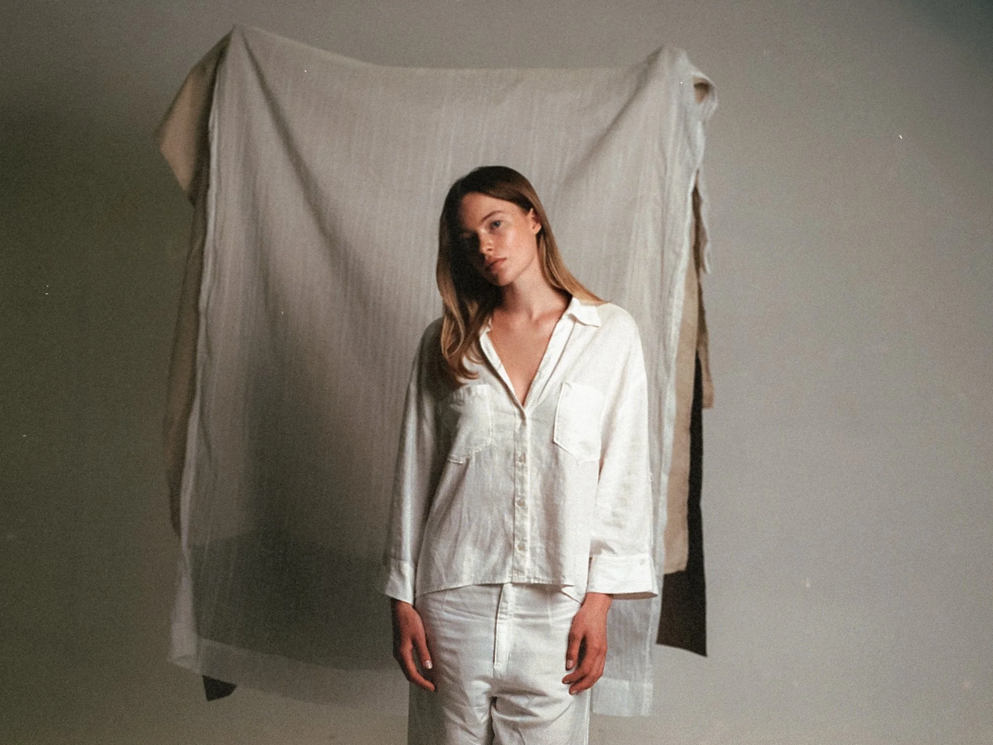 A woman with light brown hair wearing a cream-colored shirt and pants standing in front of a plain background with a fabric backdrop.