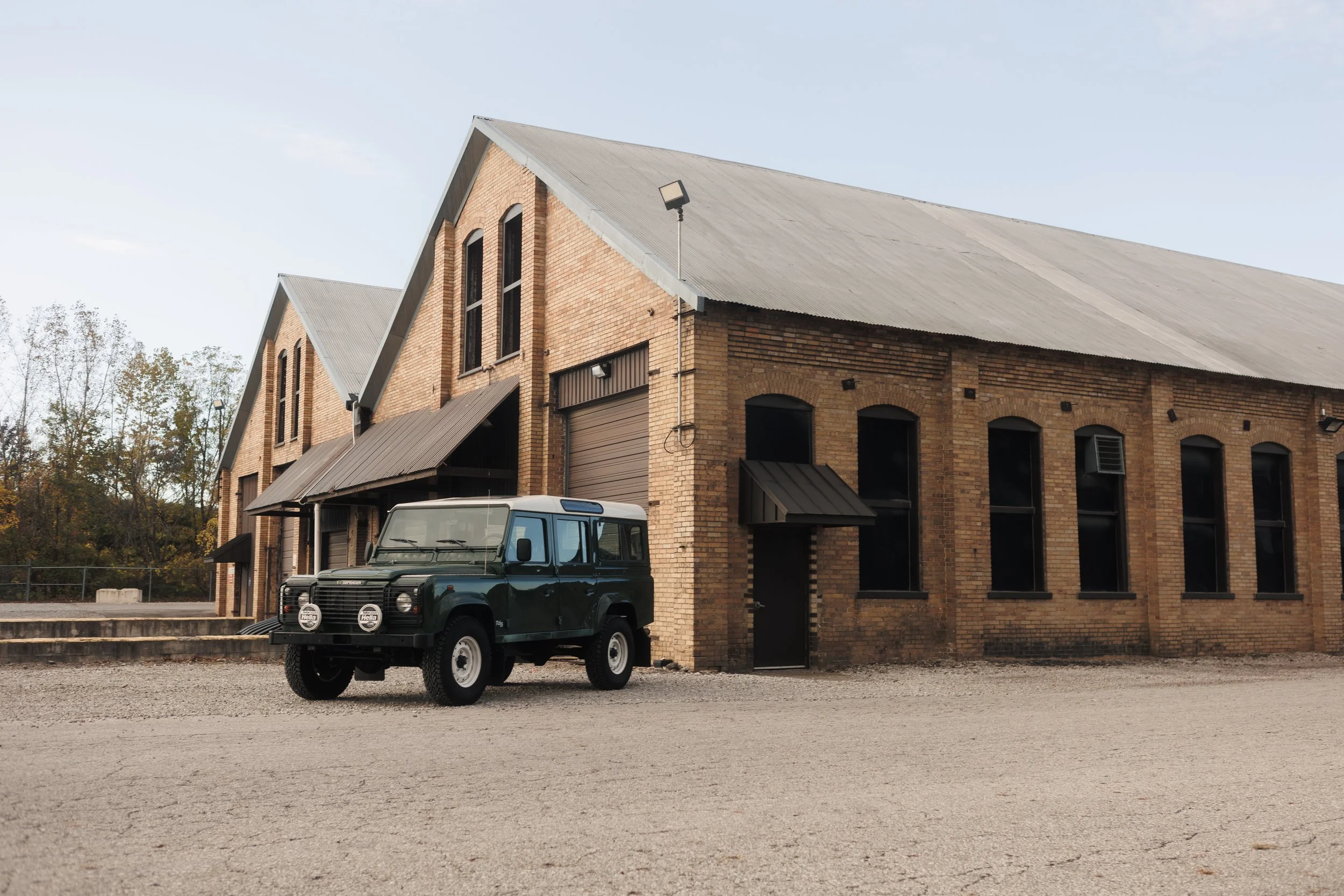 A brick building with large windows and a sloped metal roof, featuring a small awning over one door. A dark green vintage Land Rover is parked in front of the building, and there are trees in the background against a clear sky.