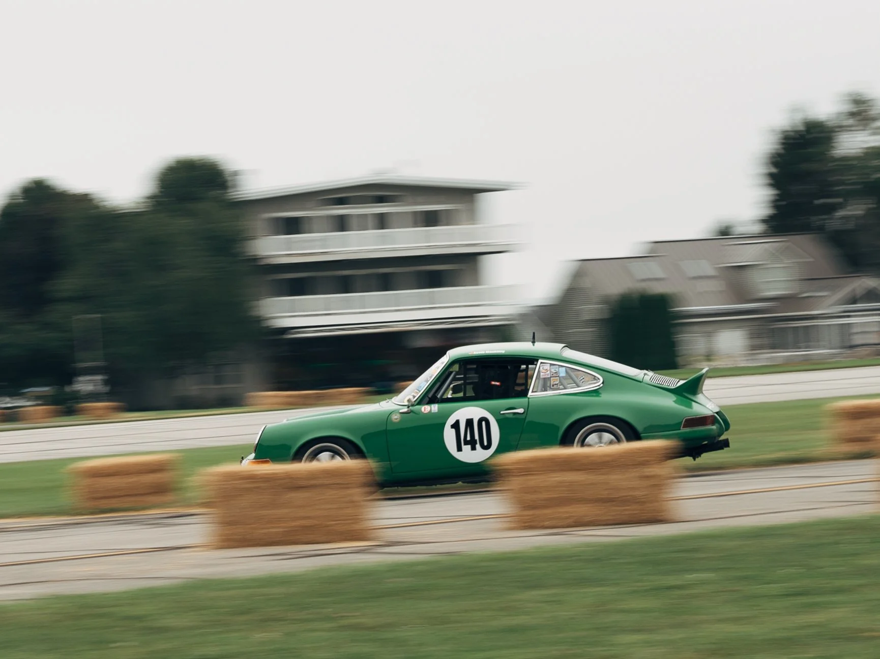 A green vintage race car with the number 140 on the door speeds along a race track, with hay bales lined up beside the track and blurred houses and trees in the background.