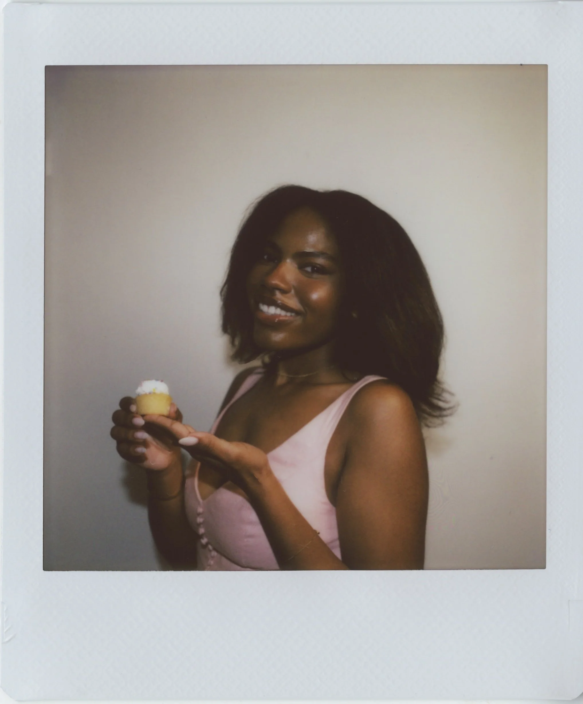 A woman with medium brown skin and long dark hair smiling, holding a small ice cream cone with white and yellow toppings, wearing a light pink sleeveless top, photographed against a plain light background.