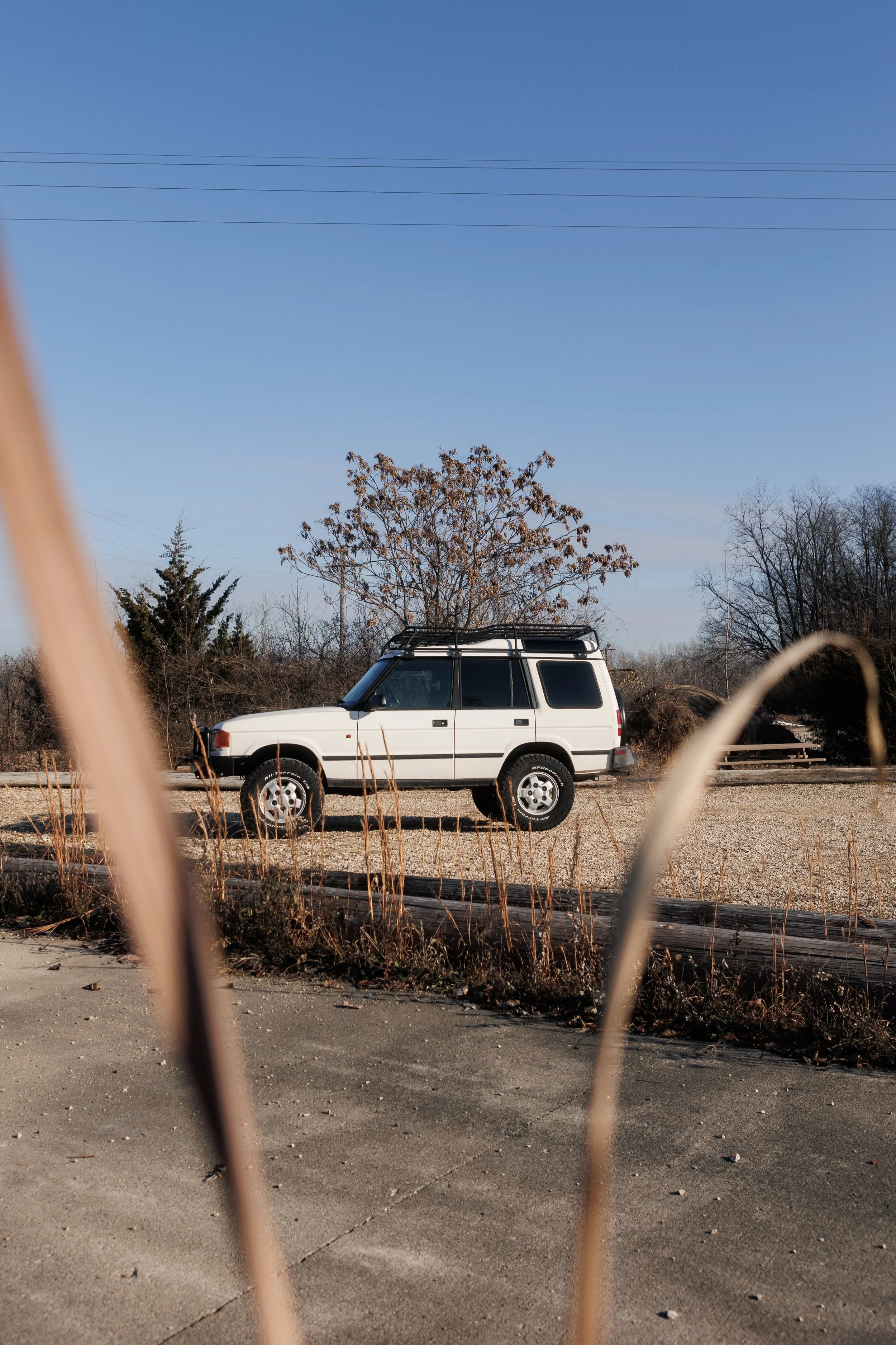 A white SUV parked on a gravel lot behind a chain-link fence, with trees and power lines in the background under a clear blue sky.