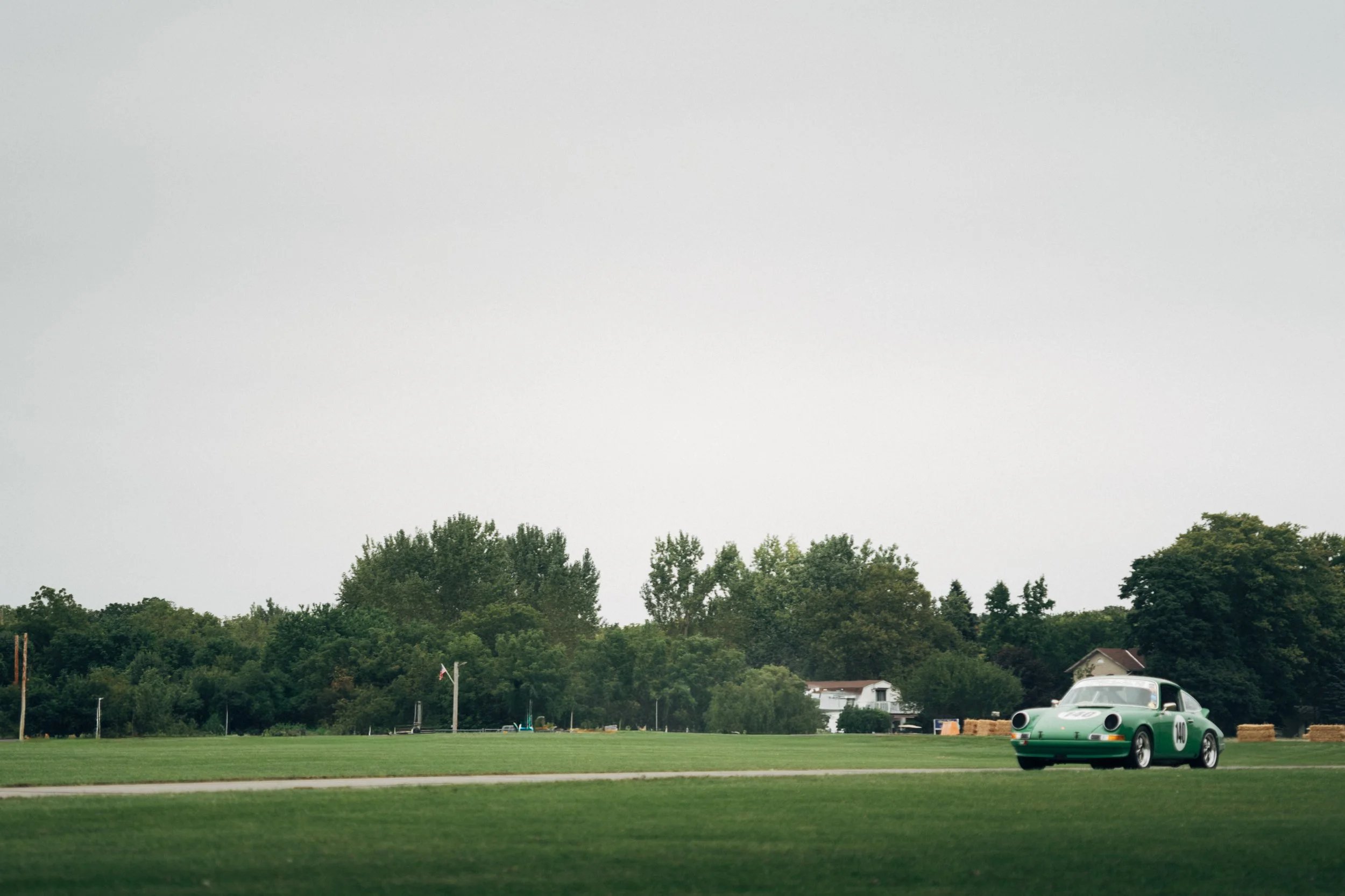 A vintage green Porsche race car parked on a grass field with trees and residential houses in the background under an overcast sky.