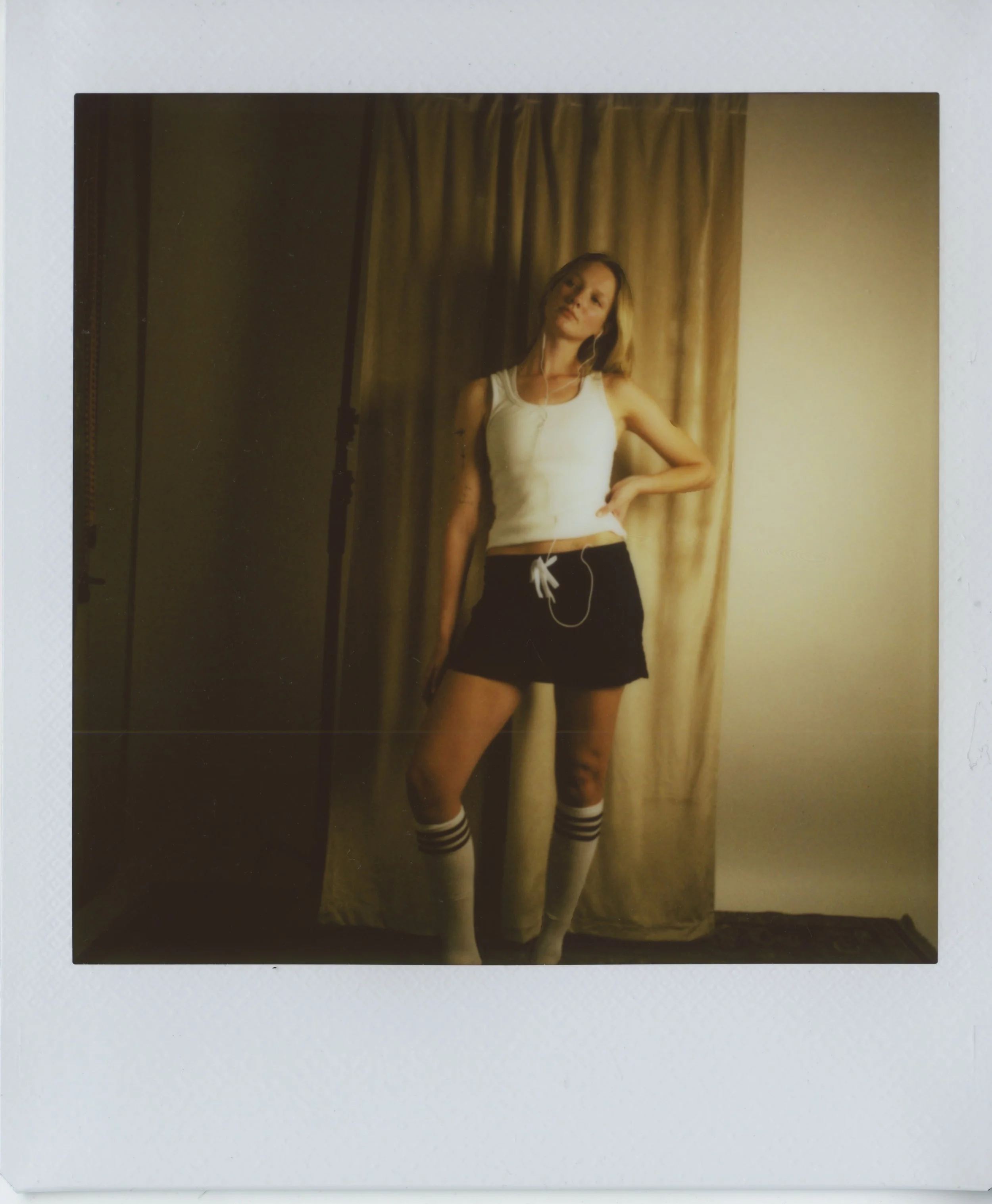 A young woman with blonde hair wearing a white tank top, black shorts, and knee-high striped socks, standing in front of beige curtains, listening to music with earphones.