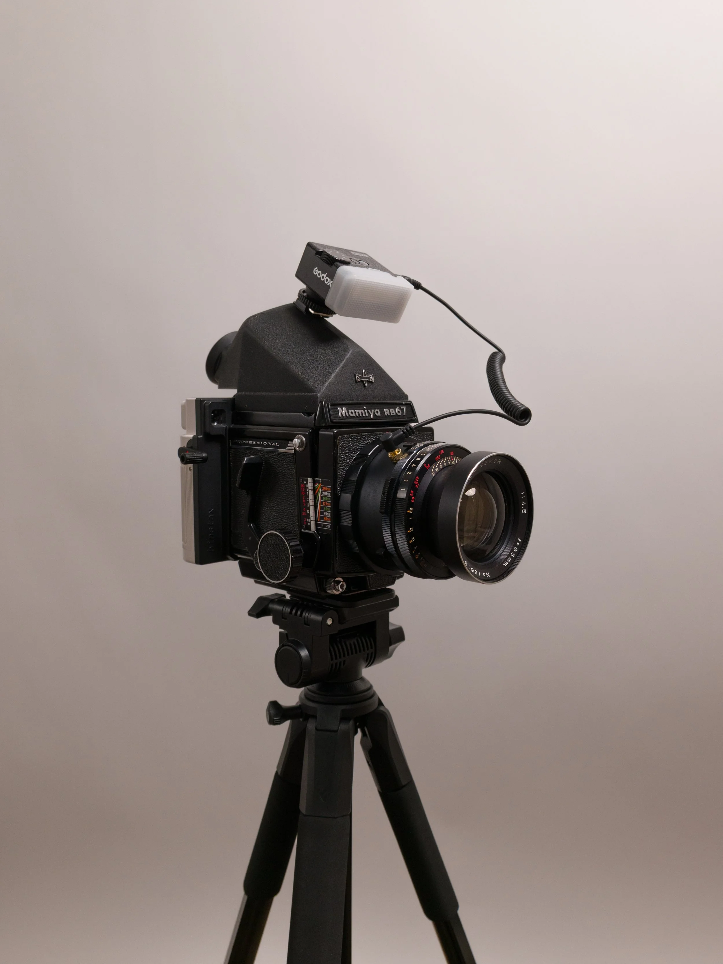 A professional film camera mounted on a tripod, with a light attached to the top, set against a plain gray background.