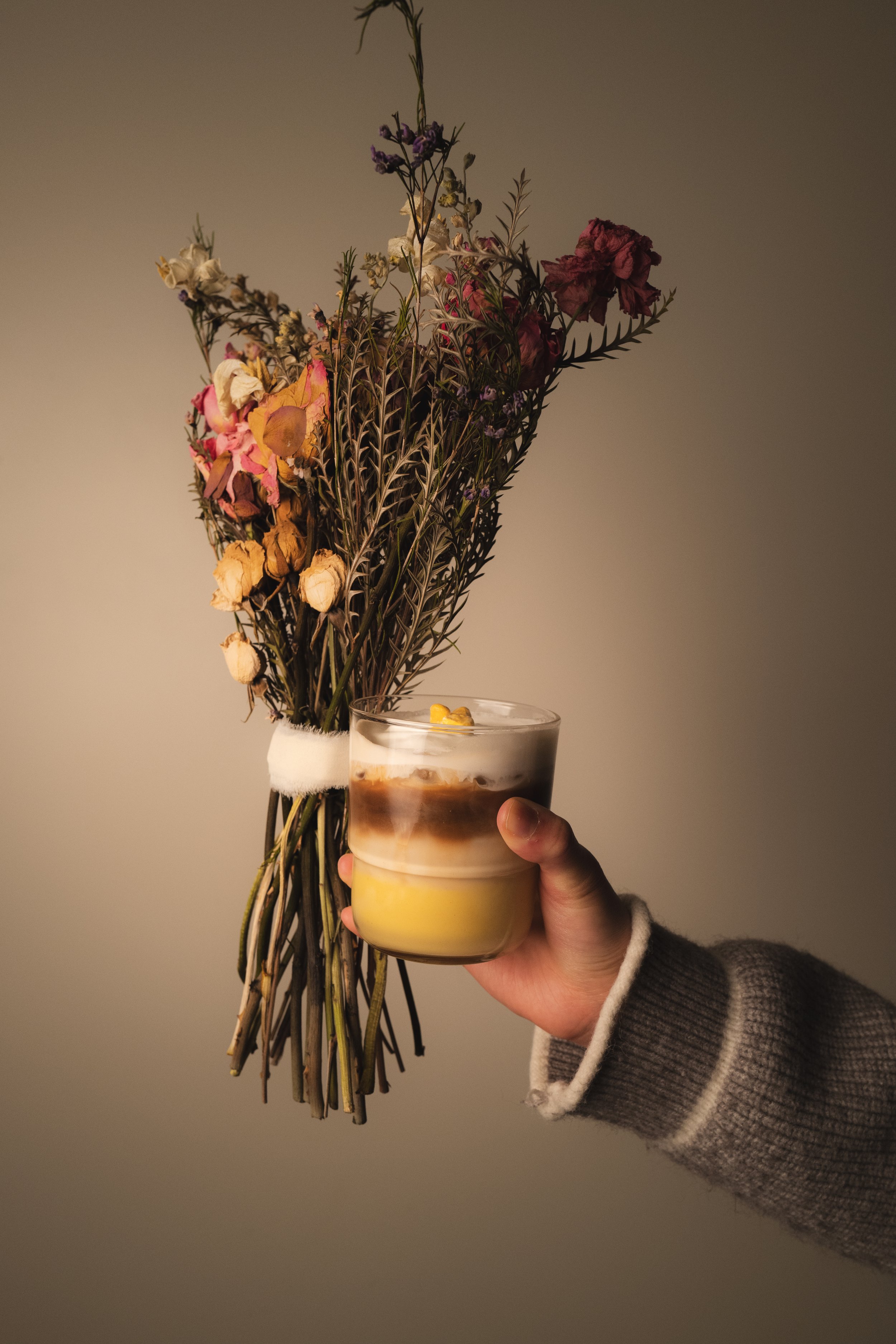 Person wearing a gray sweater with white trim is holding a layered coffee drink in front of a neutral background. Behind the drink, a bouquet of dried flowers in a white vase is visible. Columbus, Ohio, David Kim Photography