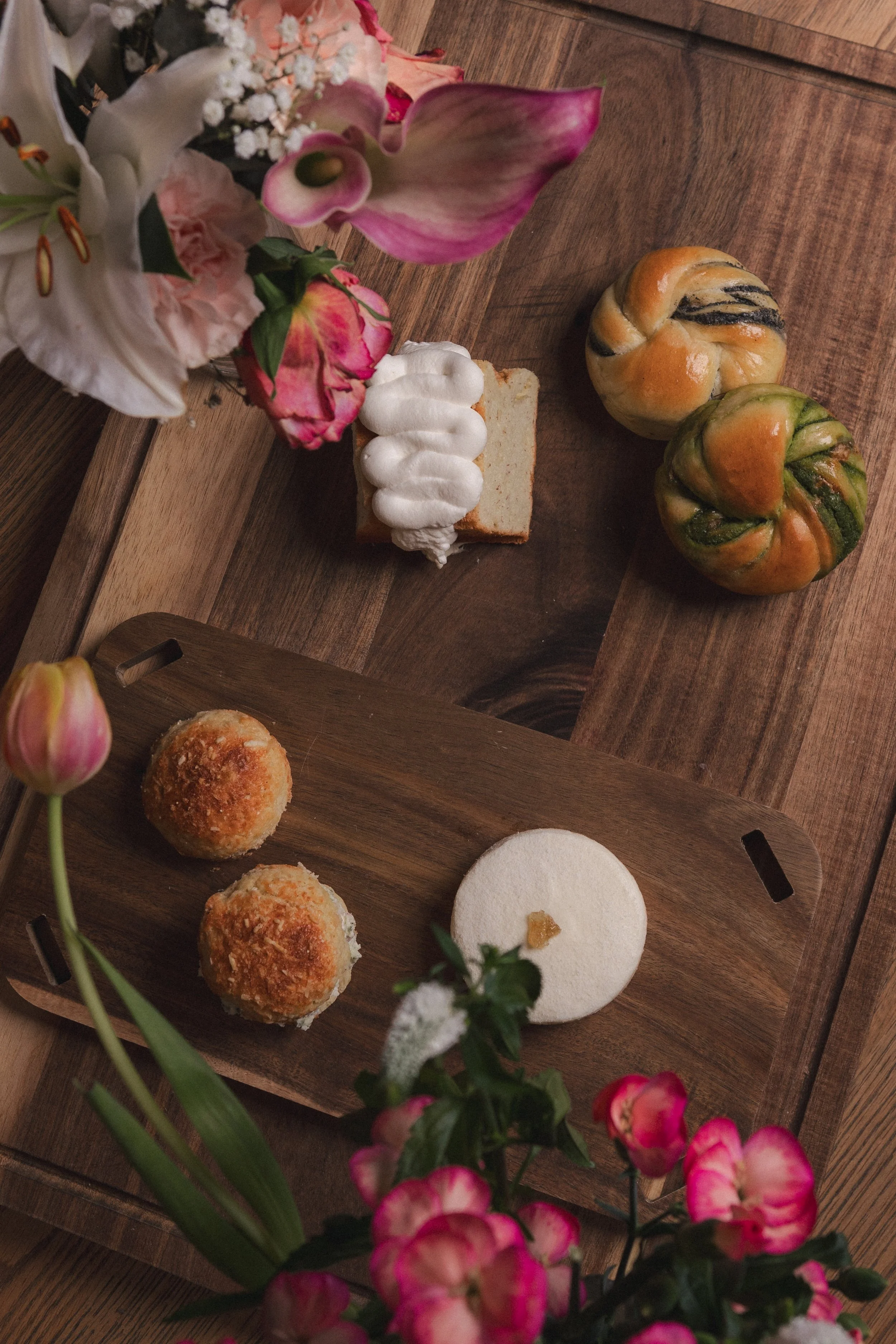A wooden tray on a table holding a variety of desserts, including a slice of cake topped with whipped cream, two round bread rolls with a crunchy coating, and a white meringue cookie. There is a bouquet of flowers with pink tulips, white lilies, and 