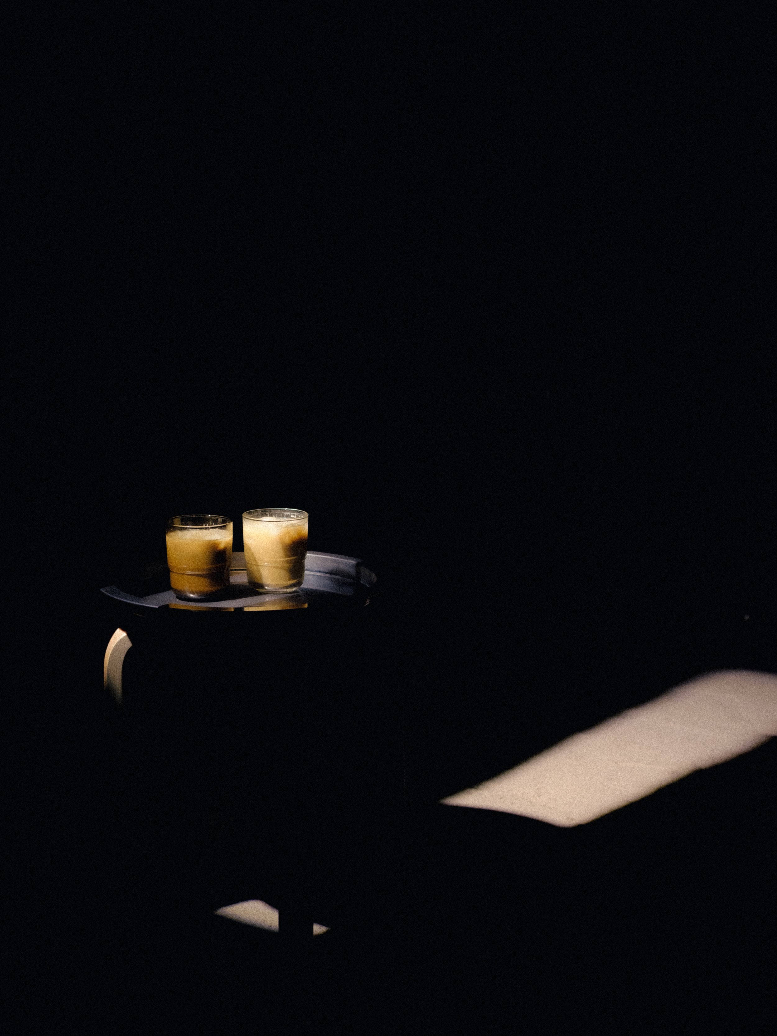 Two glasses of a light-colored beverage on a black tray, placed on a small wooden table against a dark background.
