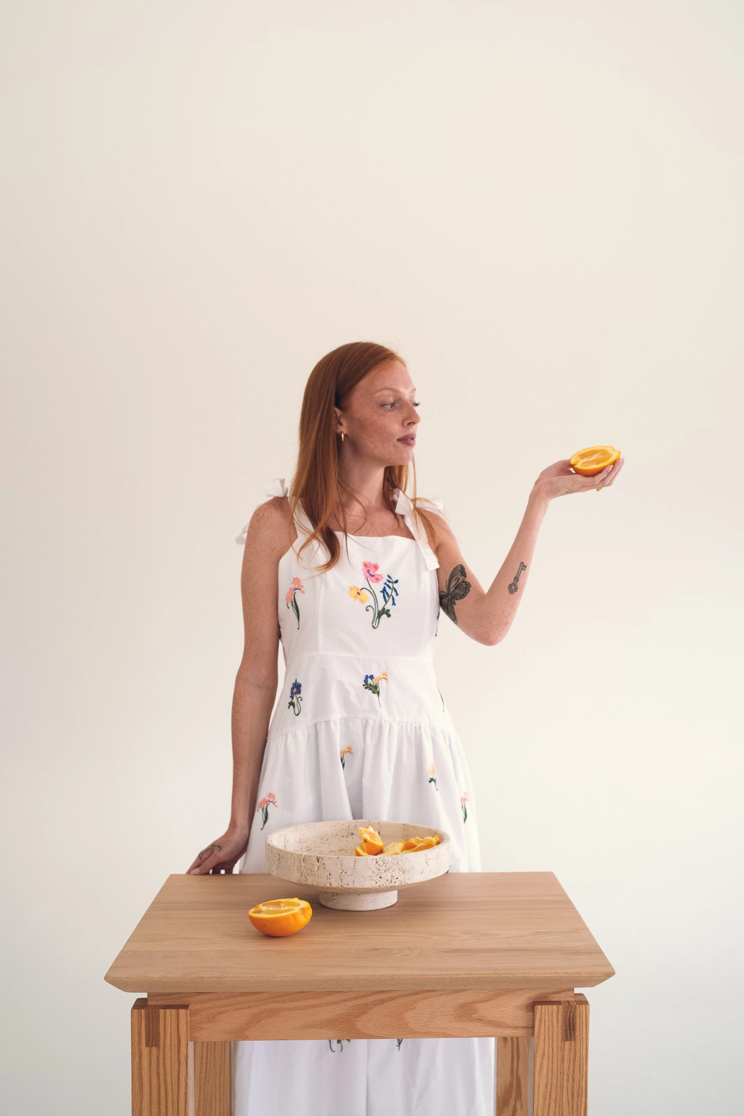 A woman with red hair wearing a white dress with colorful floral embroidery, holding a halved orange in her right hand, standing behind a wooden table with a round stone bowl filled with orange slices and a halved orange on the table. Columbus, Ohio,