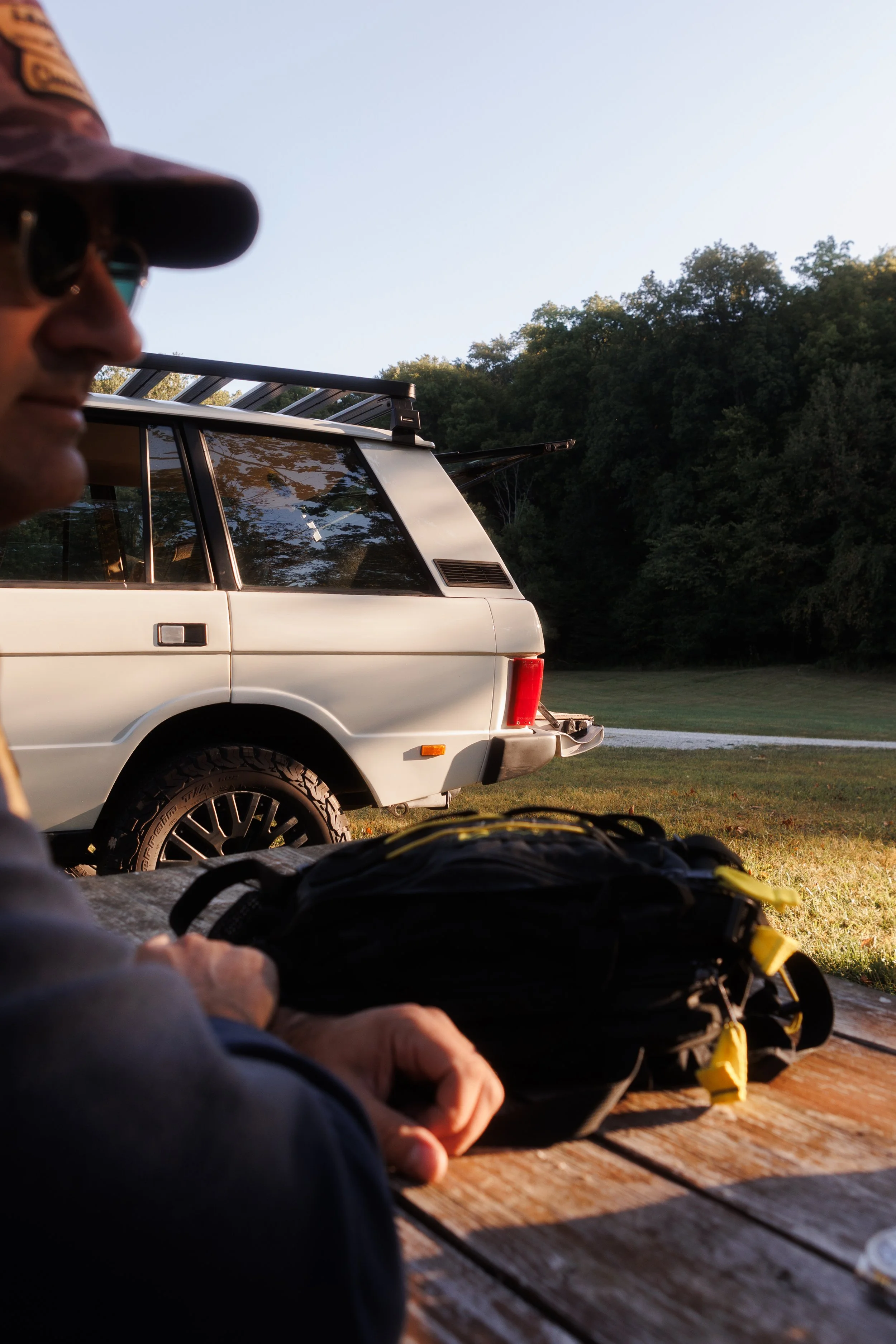 Person wearing a cap and sunglasses sitting at a wooden table outdoors, with a white vehicle and green trees in the background.
