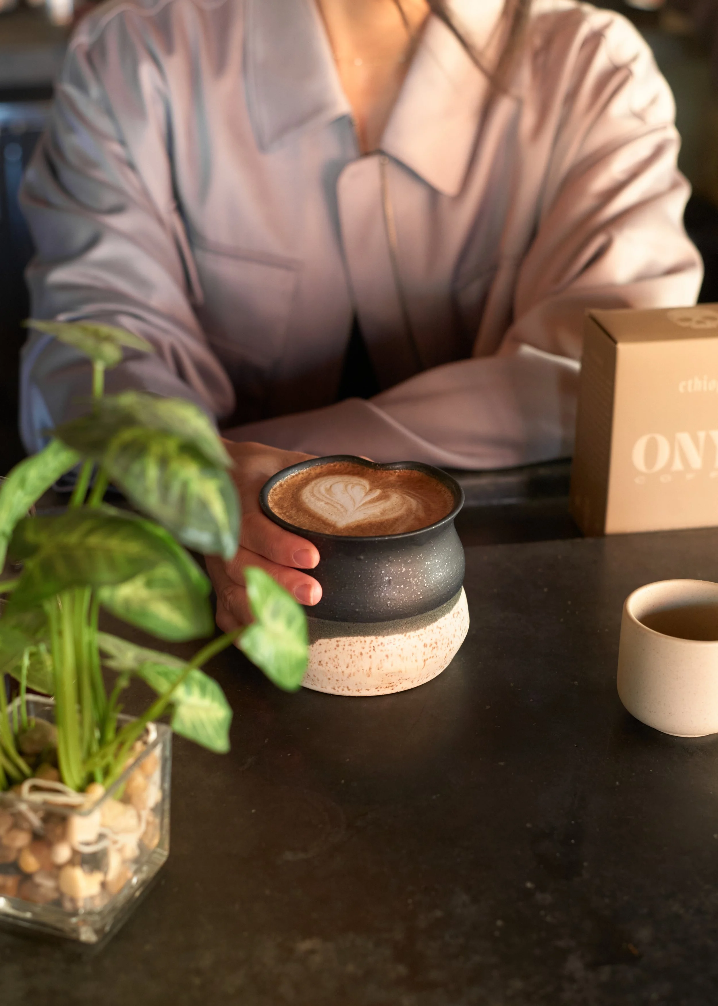 A person holding a cup of coffee with latte art, sitting at a dark table with a small potted plant, a box labeled "ONE," and a beige mug. Columbus, Ohio, David Kim Photography