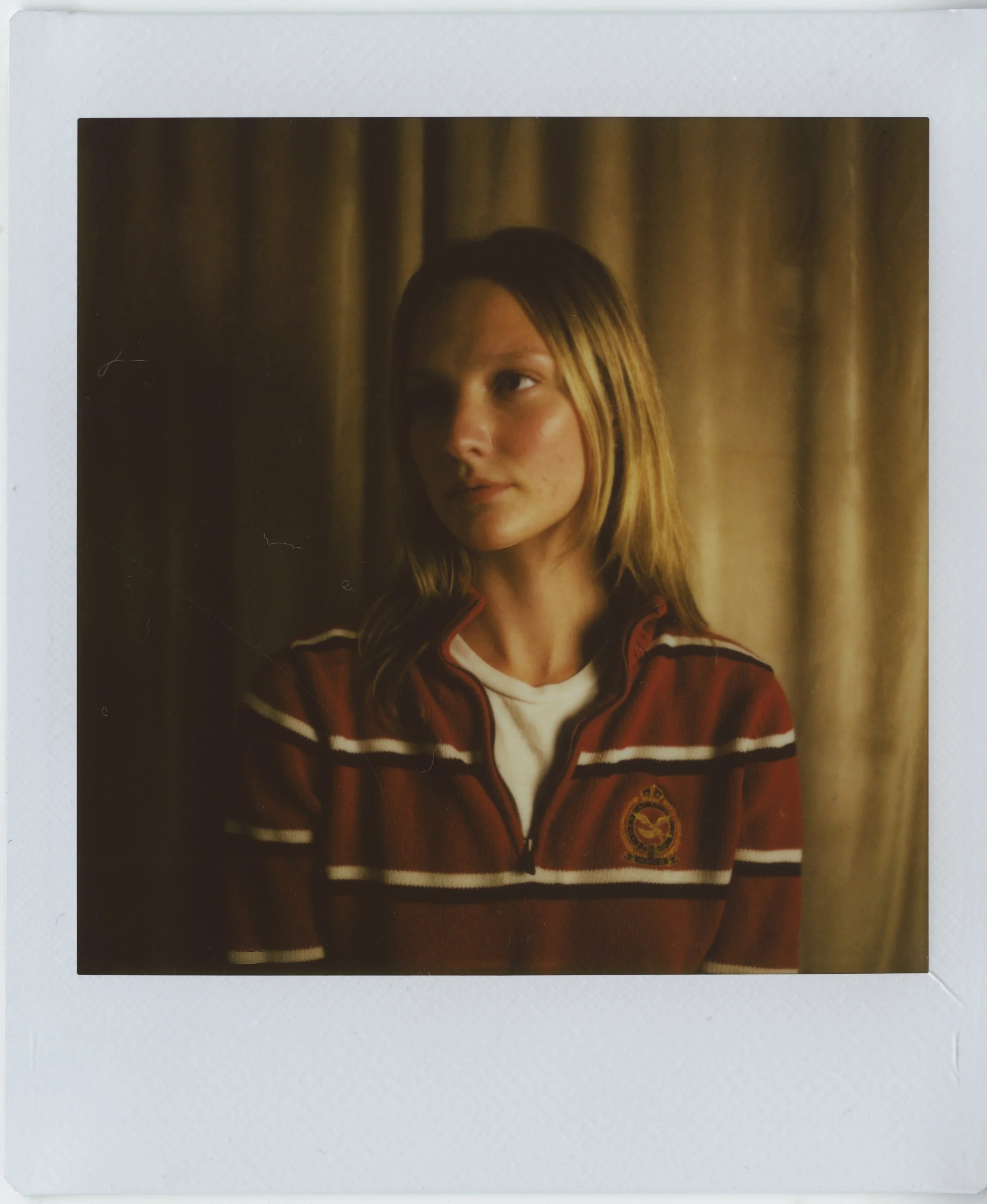 Polaroid picture of a young woman with blonde hair wearing a striped jacket with a crest on the side, with a dark background resembling a curtain.