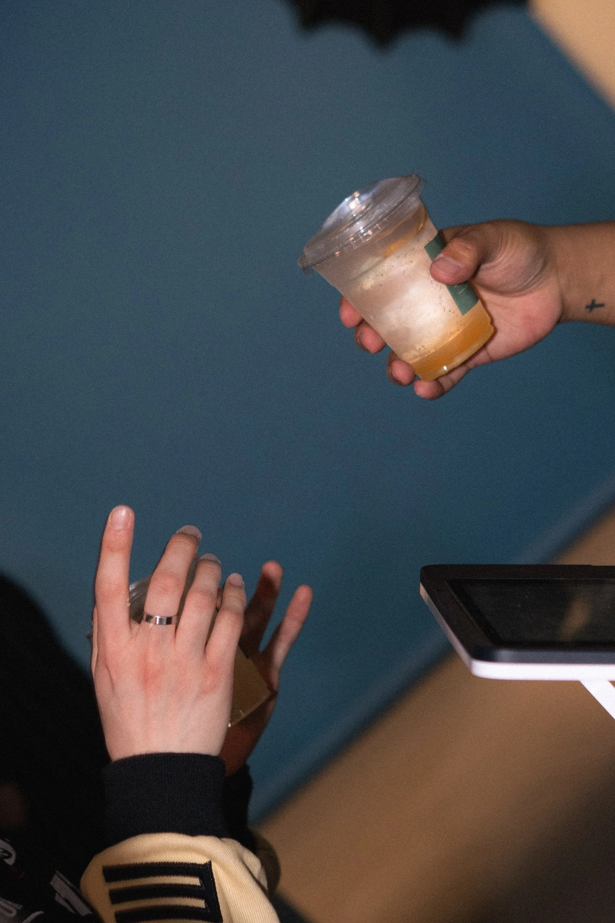 A hand holding a partially empty cup of orange juice, another hand with a finger pointing upward, a smartphone on the table, and a person's arm wearing a black and cream-colored jacket. Columbus, Ohio, David Kim Photography