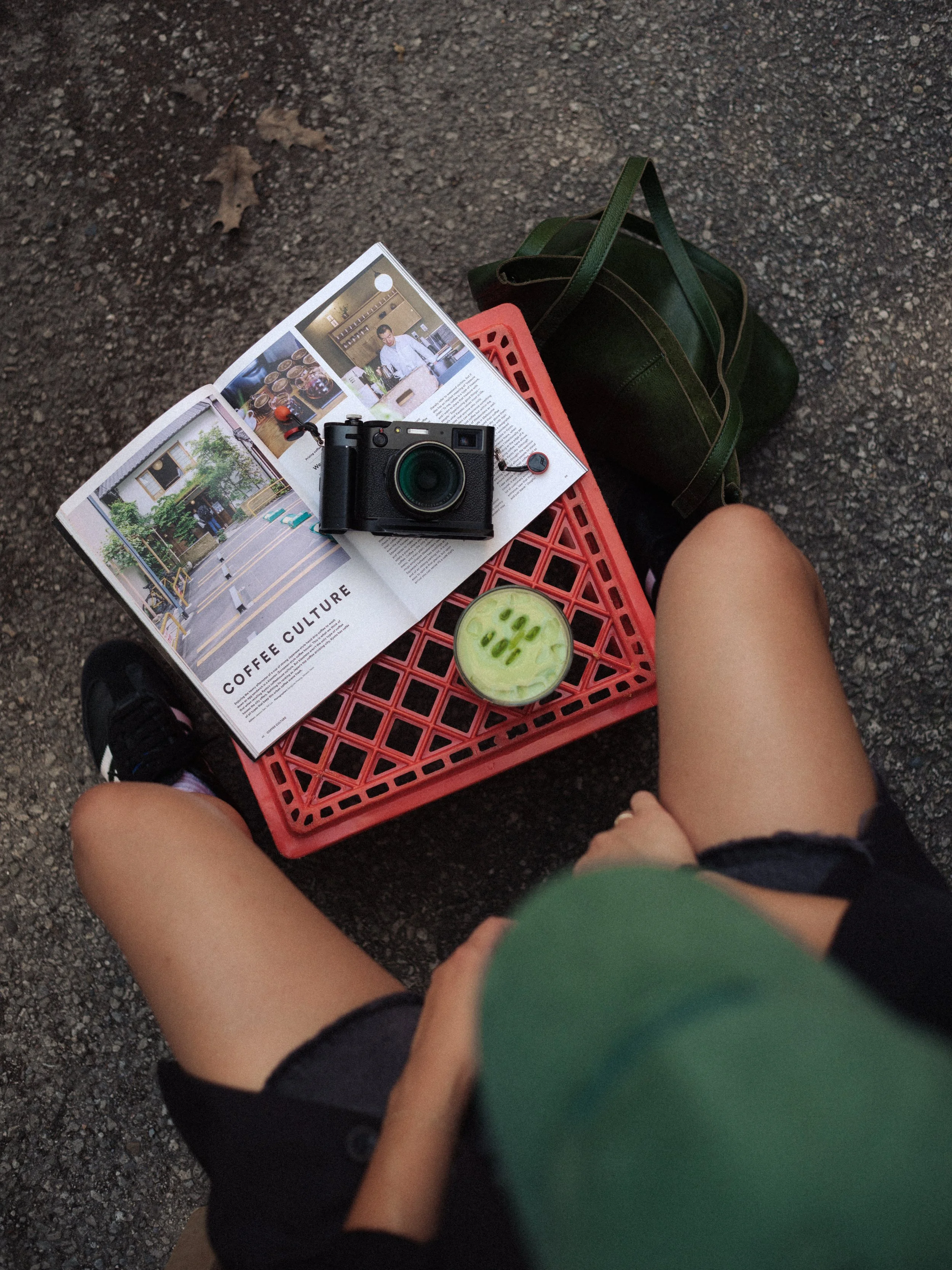 A person sitting on a red plastic crate on a gravel surface, holding a magazine open to a page with photos of a coffee shop. There is a camera placed on top of the magazine, and a green bag and a cup of matcha latte with sliced cucumbers are also vis