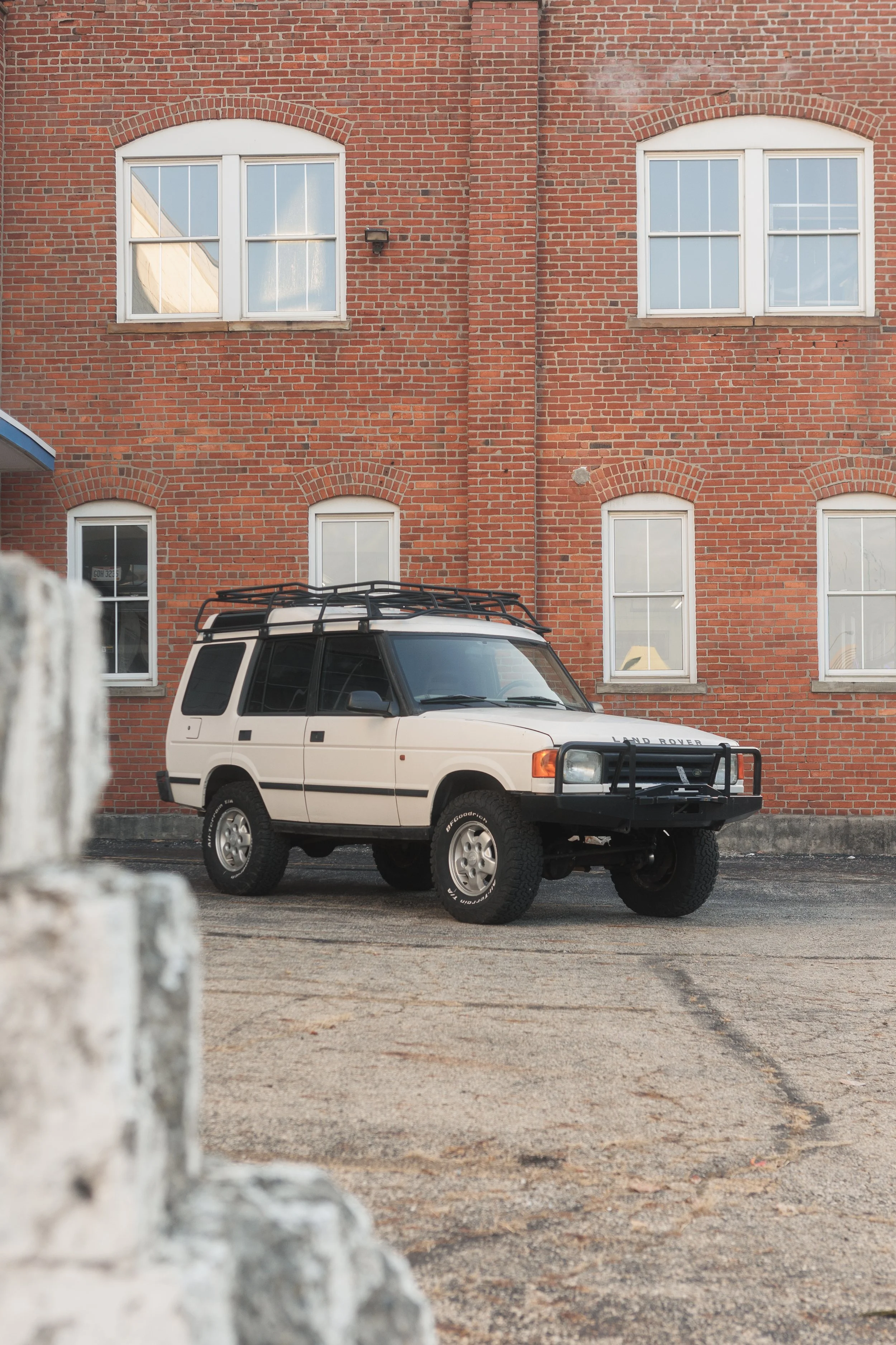 A white Land Rover SUV with a roof rack parked on a street in front of a red brick building with four windows, two on the upper floor and two on the lower floor, with reflected sky and interior items visible through the glass.