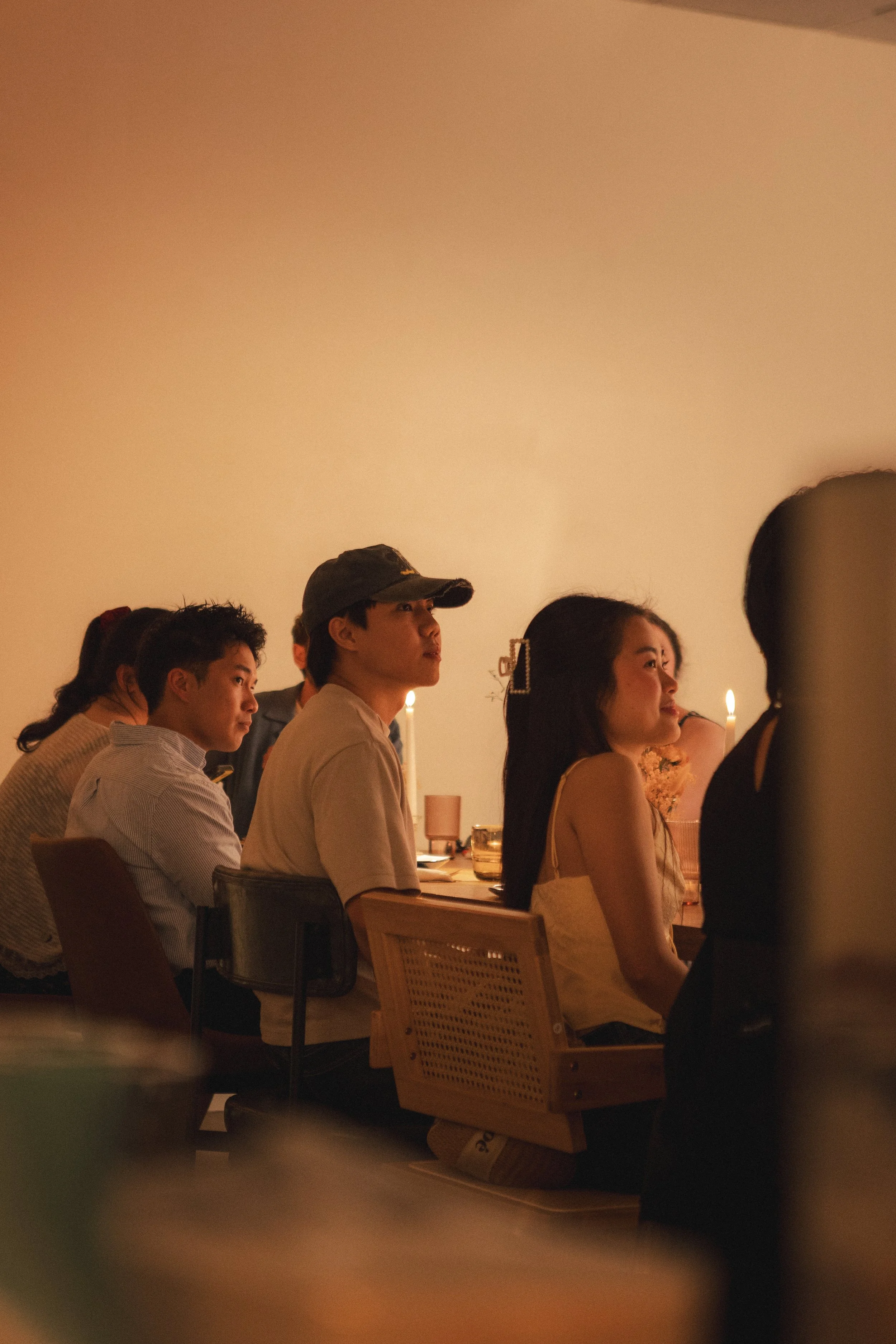 A group of people sitting at a long table, some with drinks, in a dimly lit room, possibly at a dinner or social event.