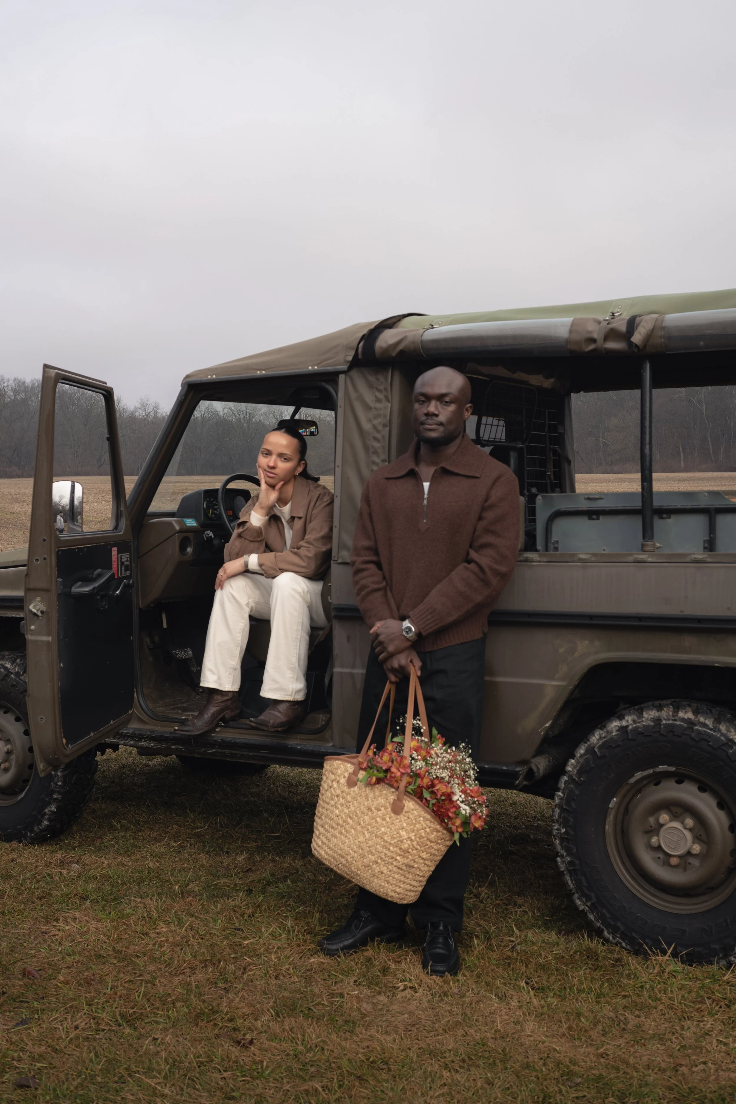 Two people, a woman sitting in the driver seat and a man standing outside, next to a military-style vehicle in a field with trees in the background. The woman is resting her chin on her hand, and the man is holding a basket of flowers. Columbus, Ohio