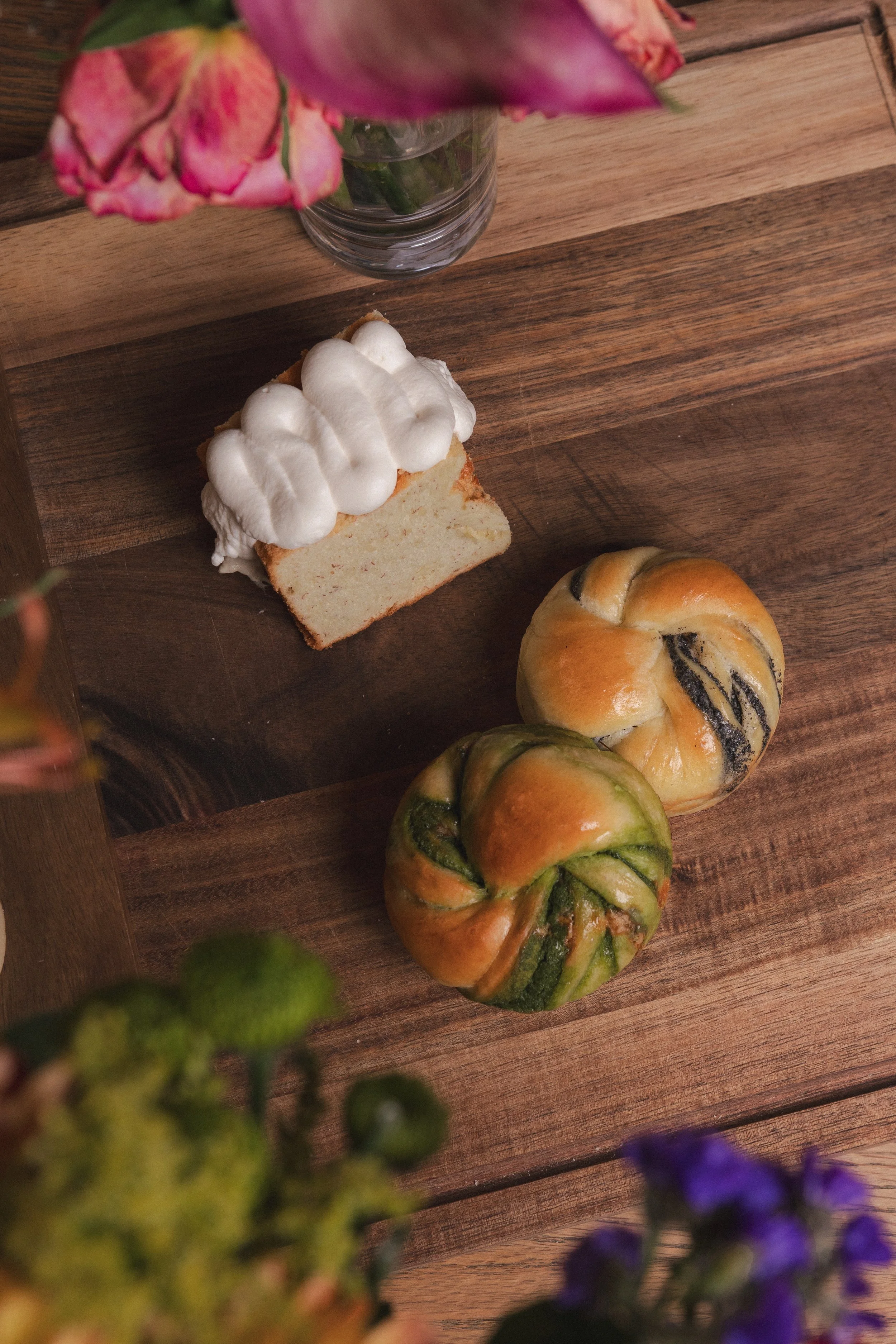 A top-down view of a wooden table with a slice of vanilla cake with white frosting, decorated with dollops of whipped topping, a round pumpkin, and a floral arrangement with pink and purple flowers in the background.