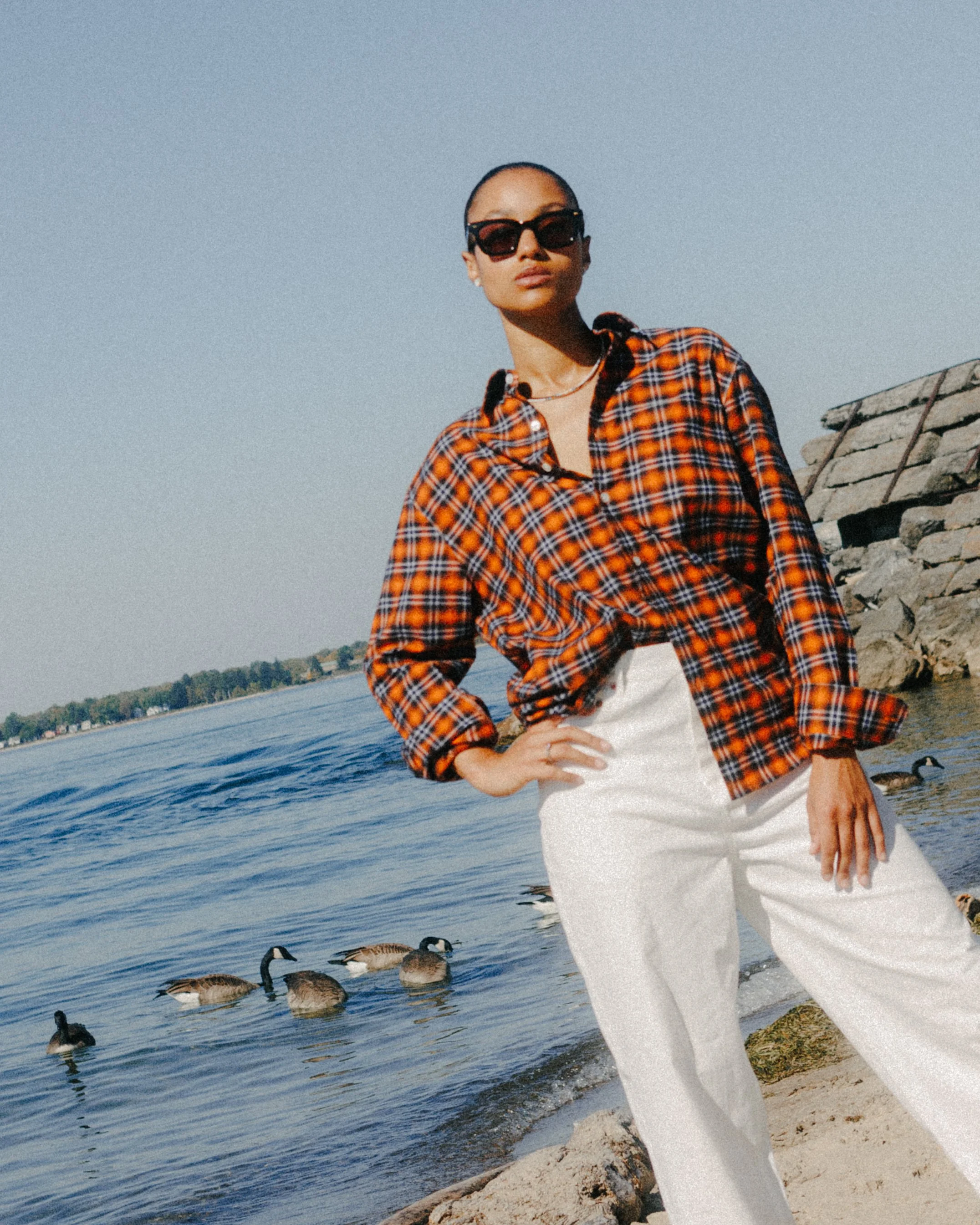 A woman standing by a riverbank with ducks swimming in the water behind her. She is wearing sunglasses, a red and black plaid shirt, and white pants, with her hand on her hip and the other hanging by her side.